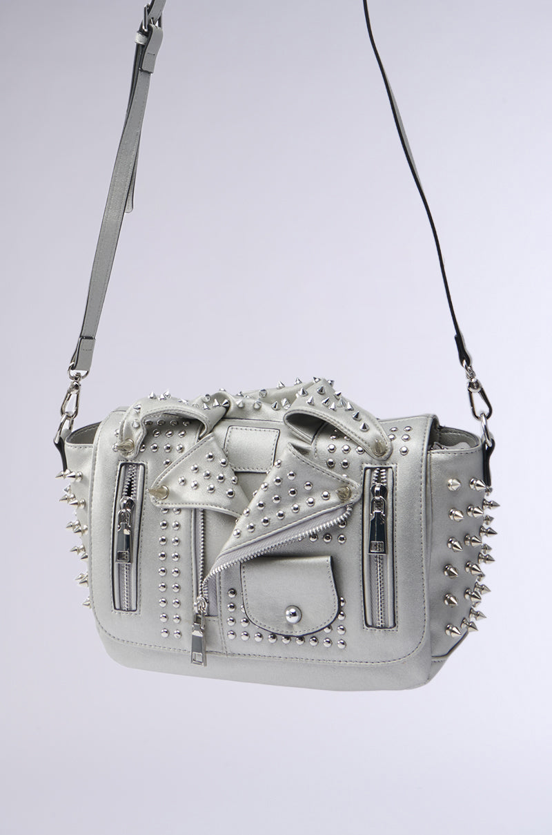 The MOTO OF YOUR DREAMS PURSE is a stylish metallic silver handbag with metal studs and zippers, mimicking a moto jacket. It boasts two front zipper pockets, decorative buttons, and an adjustable shoulder strap for an edgy, fashionable appearance.
