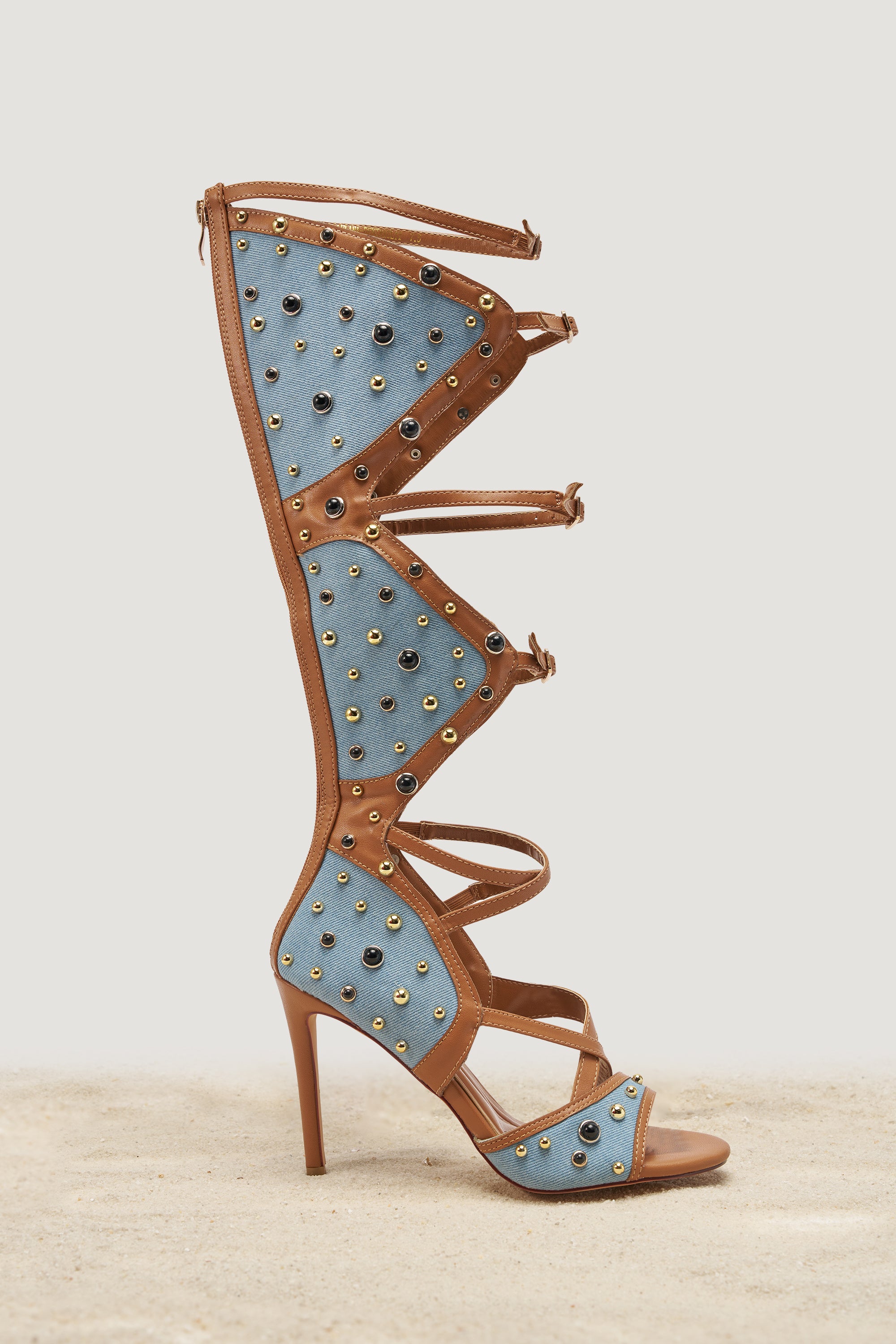 The AKIRA GOLD LULANA DENIM METAL STUDDED GLADIATOR STILETTO SANDAL features knee-high denim panels, tan leather straps, and gold/silver studs—an eye-catching, open-toe statement shoe for festivals, displayed on a neutral background. 1