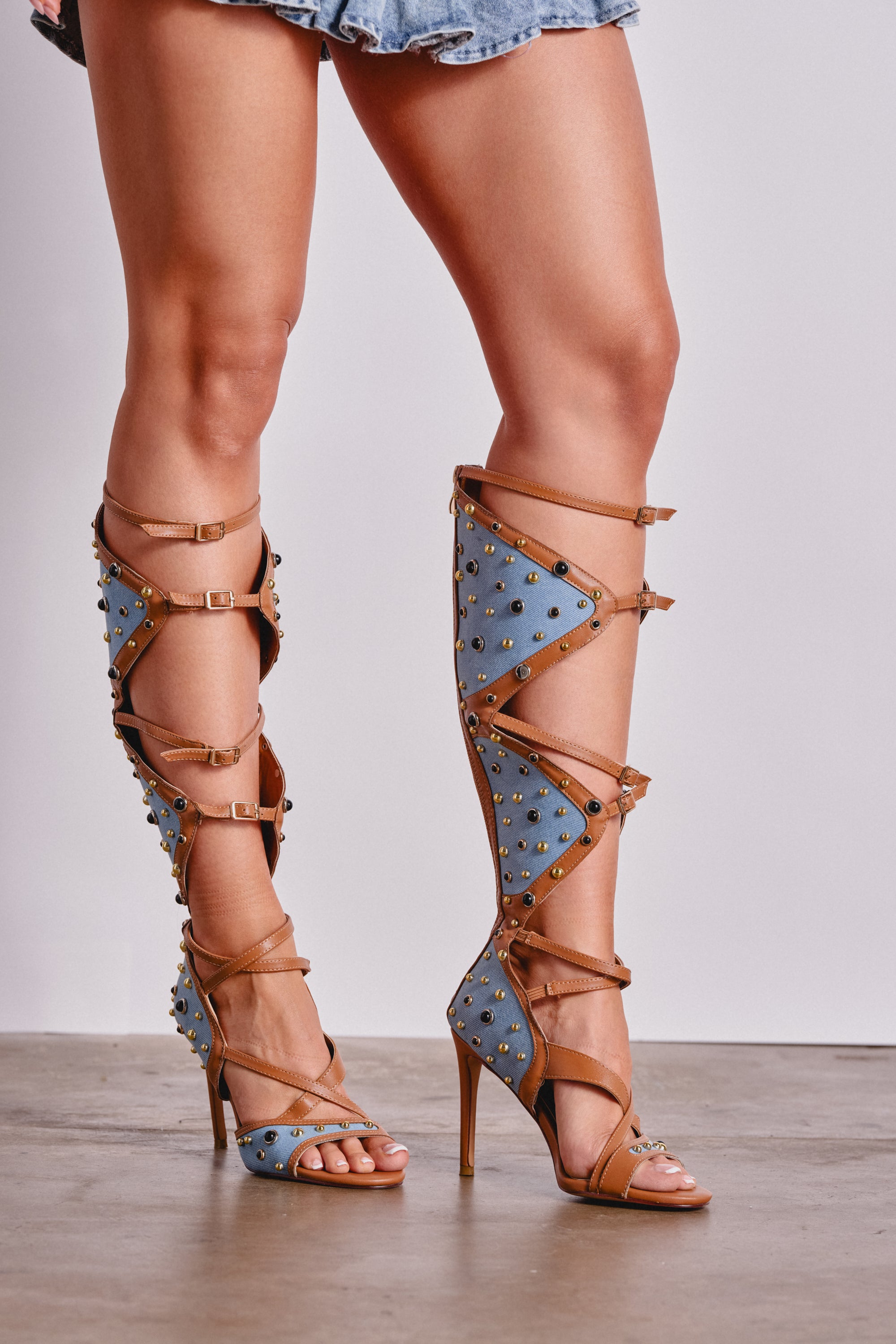 A person wears the AKIRA GOLD LULANA DENIM METAL STUDDED GLADIATOR STILETTO SANDAL—knee-high, open-toe sandals with blue panels, brown straps, gold studs, and multiple buckles—paired with a short frayed denim skirt. 0