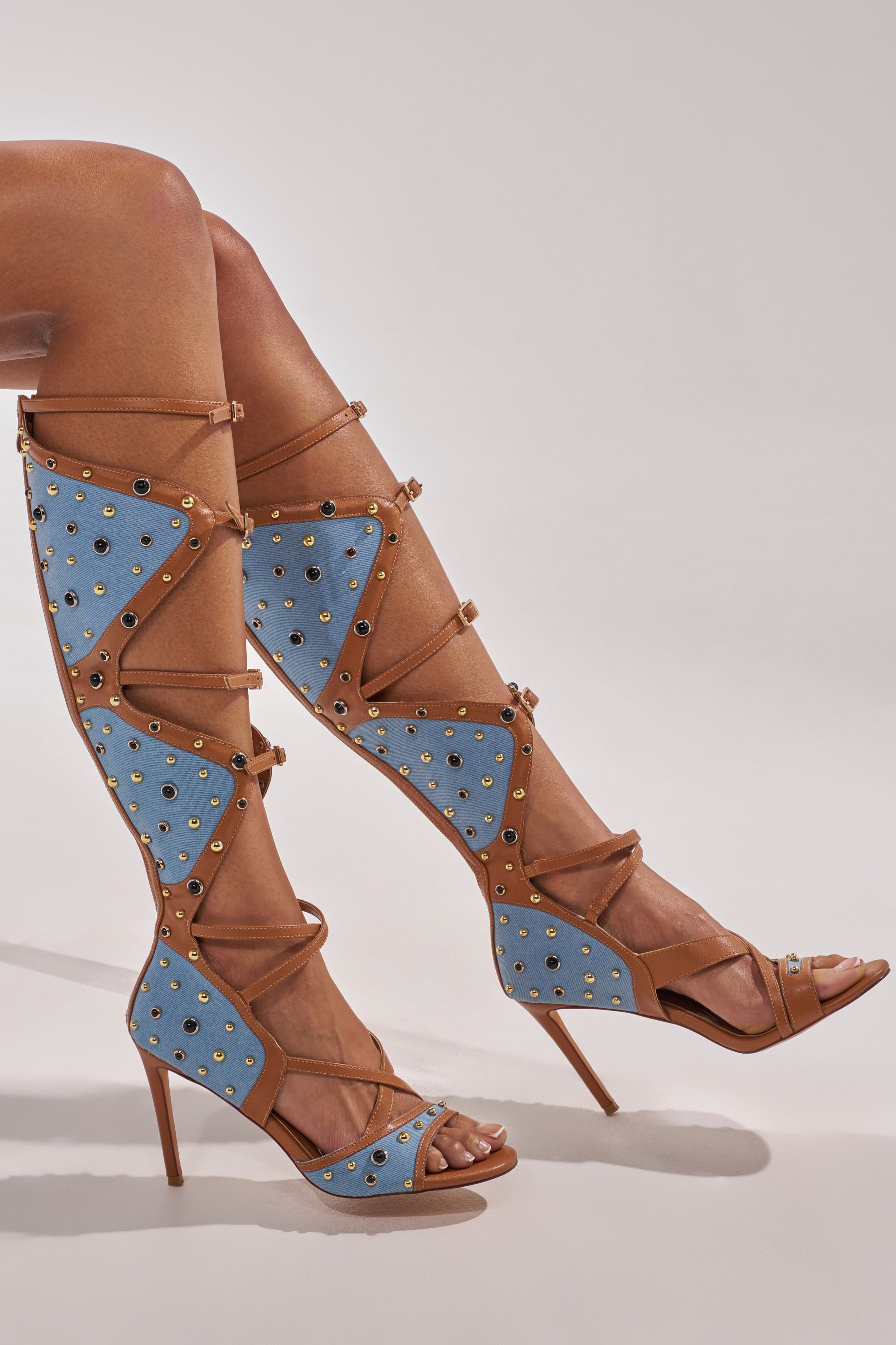 Wearing the AKIRA GOLD LULANA DENIM METAL STUDDED GLADIATOR STILETTO SANDAL, featuring knee-high blue denim panels, brown straps, and gold studs, a person sits with legs crossed against a light background. 3