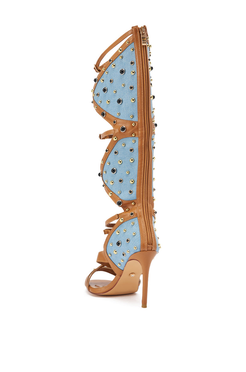 The AKIRA GOLD LULANA DENIM METAL STUDDED GLADIATOR STILETTO SANDAL features a gladiator style with denim panels, gold and silver metal studs, butterfly wing details, tan leather trim and straps, a back zip closure, and a bold stiletto heel. 7