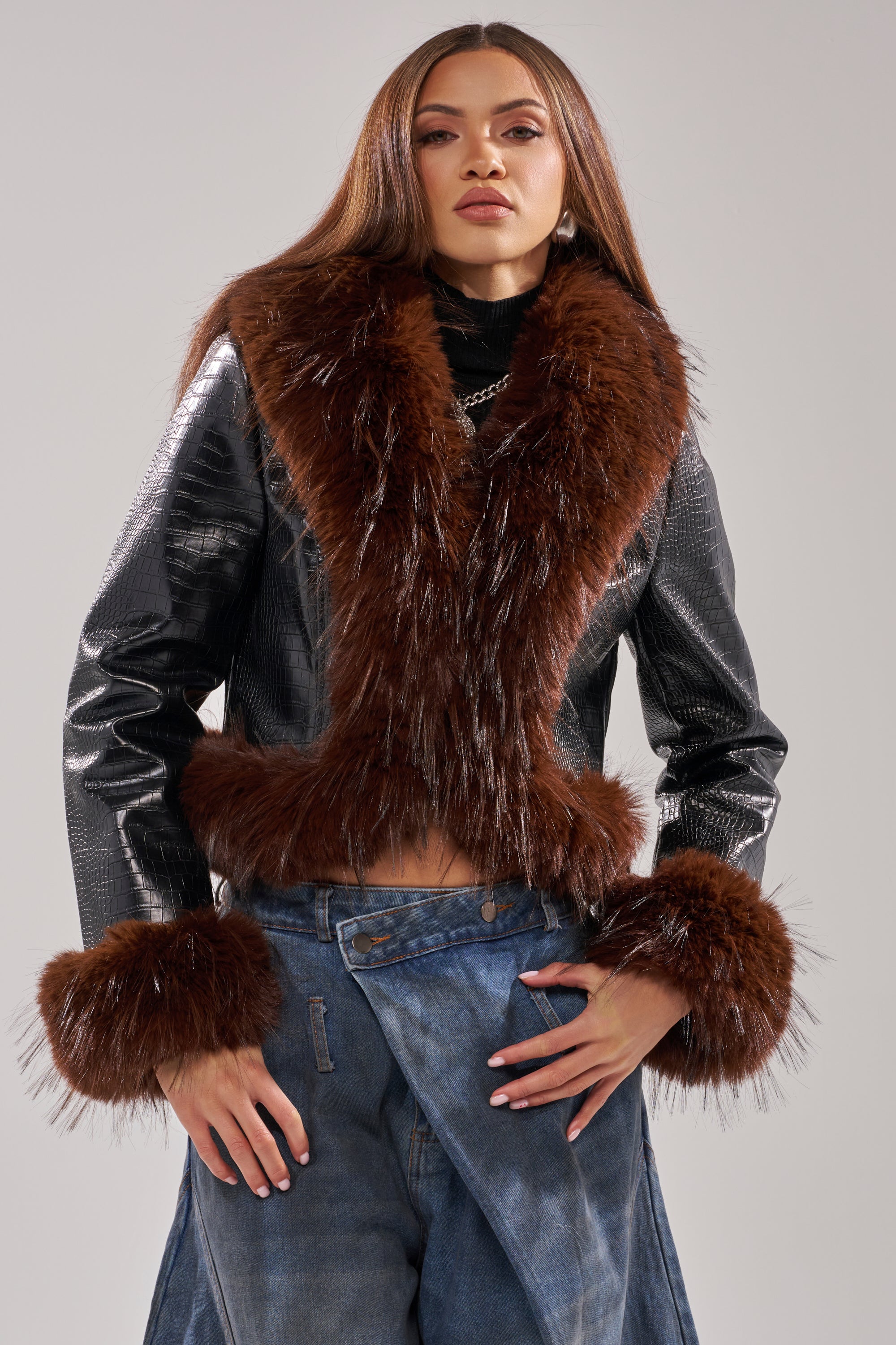 A woman poses confidently in the LUNNA FUR TRIM MOTO IN BLACK CROC, layered over a black top and paired with high-waisted, draped blue jeans against a plain light background.