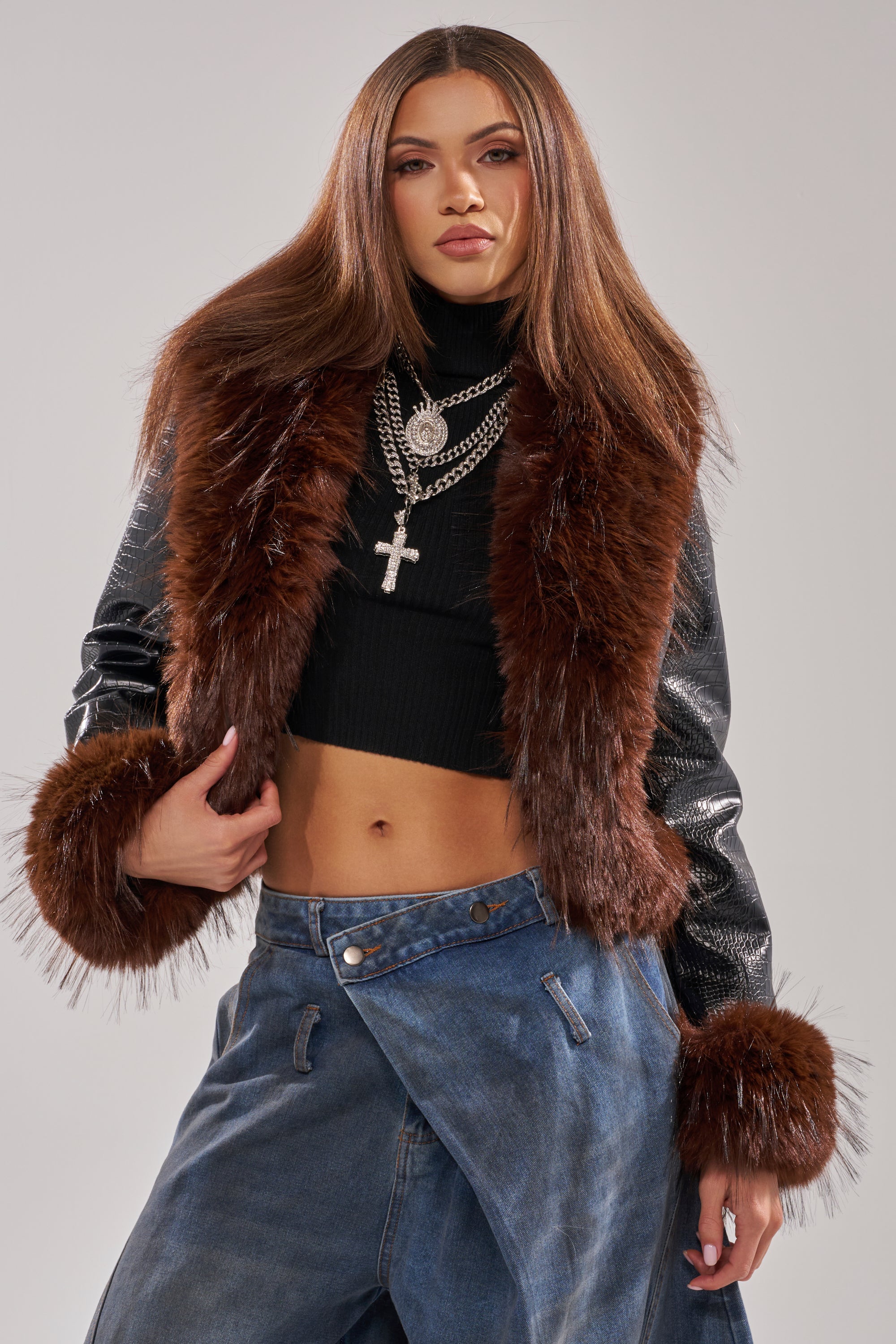 A woman with straight brown hair wears a black crop top, the LUNNA FUR TRIM MOTO IN BLACK CROC with brown fur trim, layered silver cross necklaces, and loose asymmetric blue jeans against a plain background.