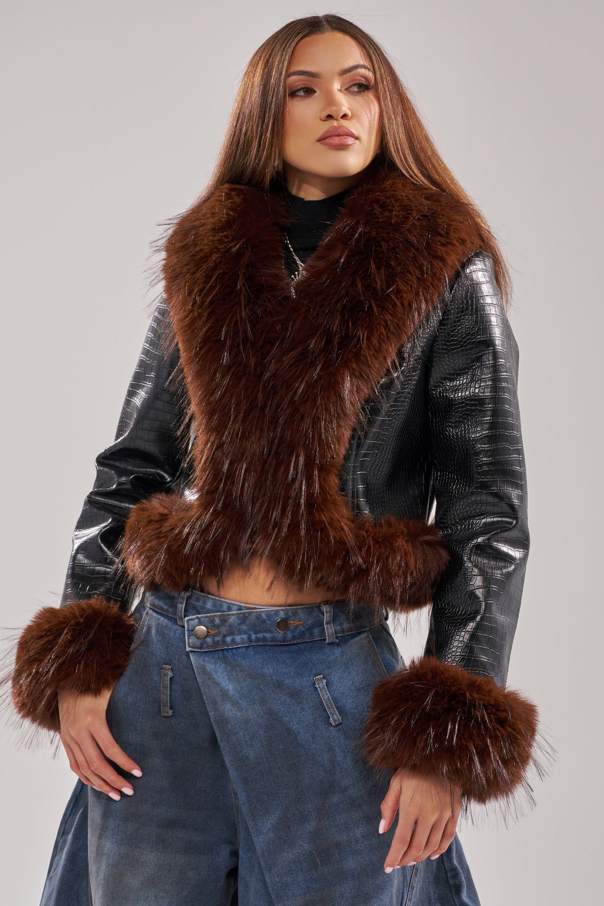 A woman models the LUNNA FUR TRIM MOTO IN BLACK CROC, styled with wide-legged blue jeans. She stands confidently against a neutral background.