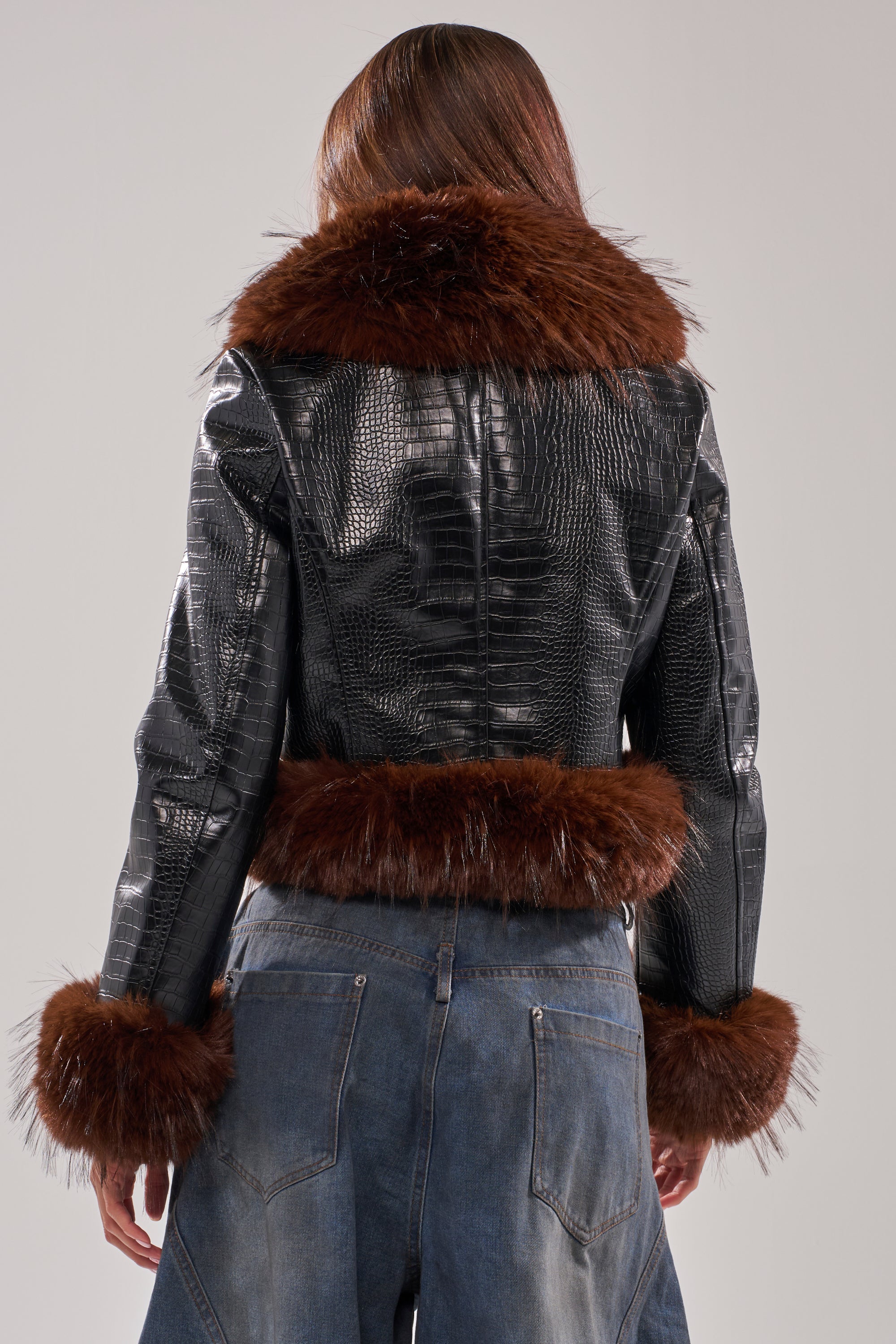 Model wears the LUNNA FUR TRIM MOTO IN BLACK CROC, featuring brown fur on the collar, cuffs, and hem, paired with high-waisted blue jeans. The look is viewed from the back.