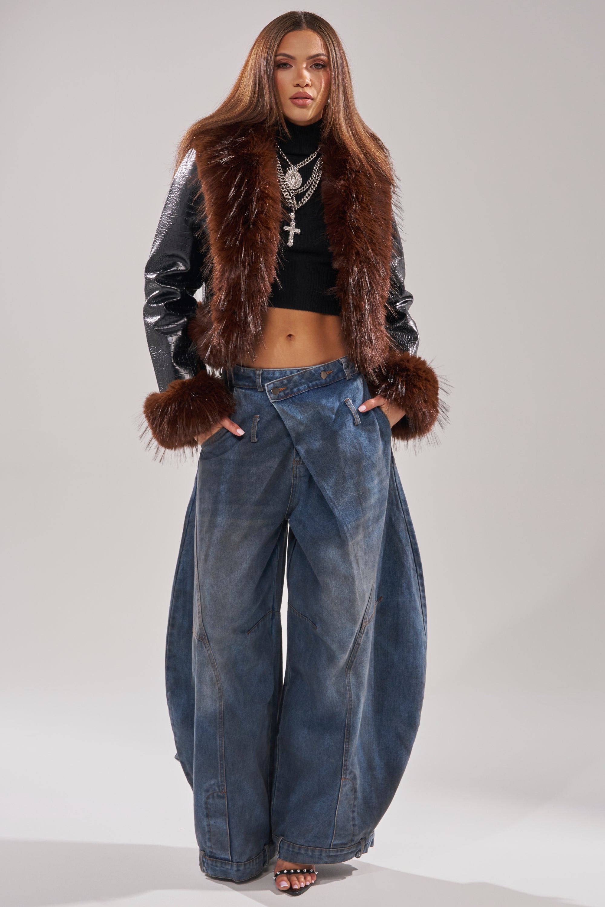 A woman stands confidently in wide-leg denim, a black crop top, and the LUNNA FUR TRIM MOTO IN BLACK CROC, layered with necklaces and open-toe heels against a plain light background.