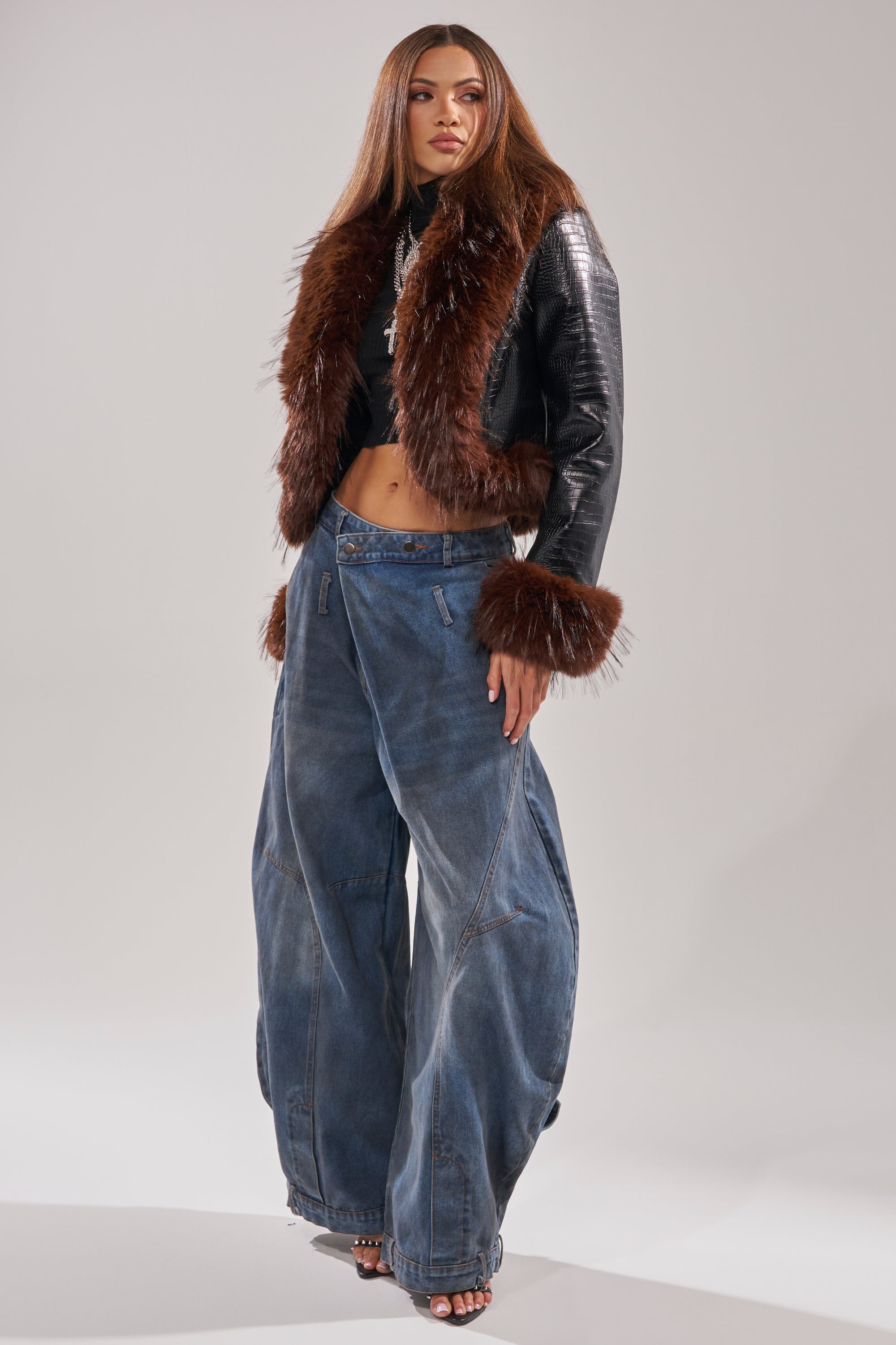A woman poses confidently against a plain background in the LUNNA FUR TRIM MOTO IN BLACK CROC, styled with wide-leg blue jeans, a black crop top, open-toe heels, and her long straight hair complementing the look.