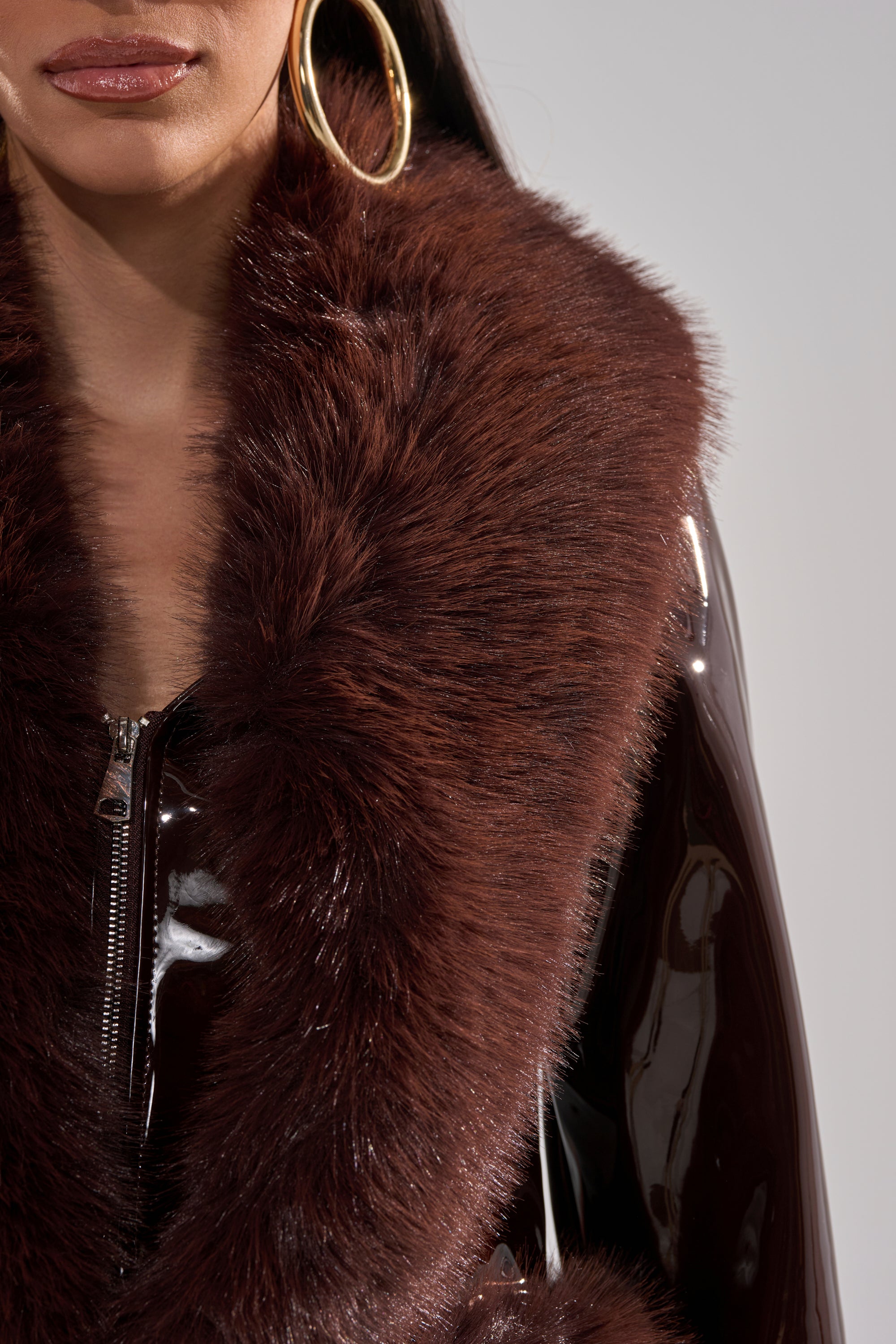 A woman wears the LUNNA FUR TRIM MOTO IN PATENT—a glossy faux leather jacket with a wide, fluffy faux fur collar—and large gold hoop earrings, shown from her nose to midsection.