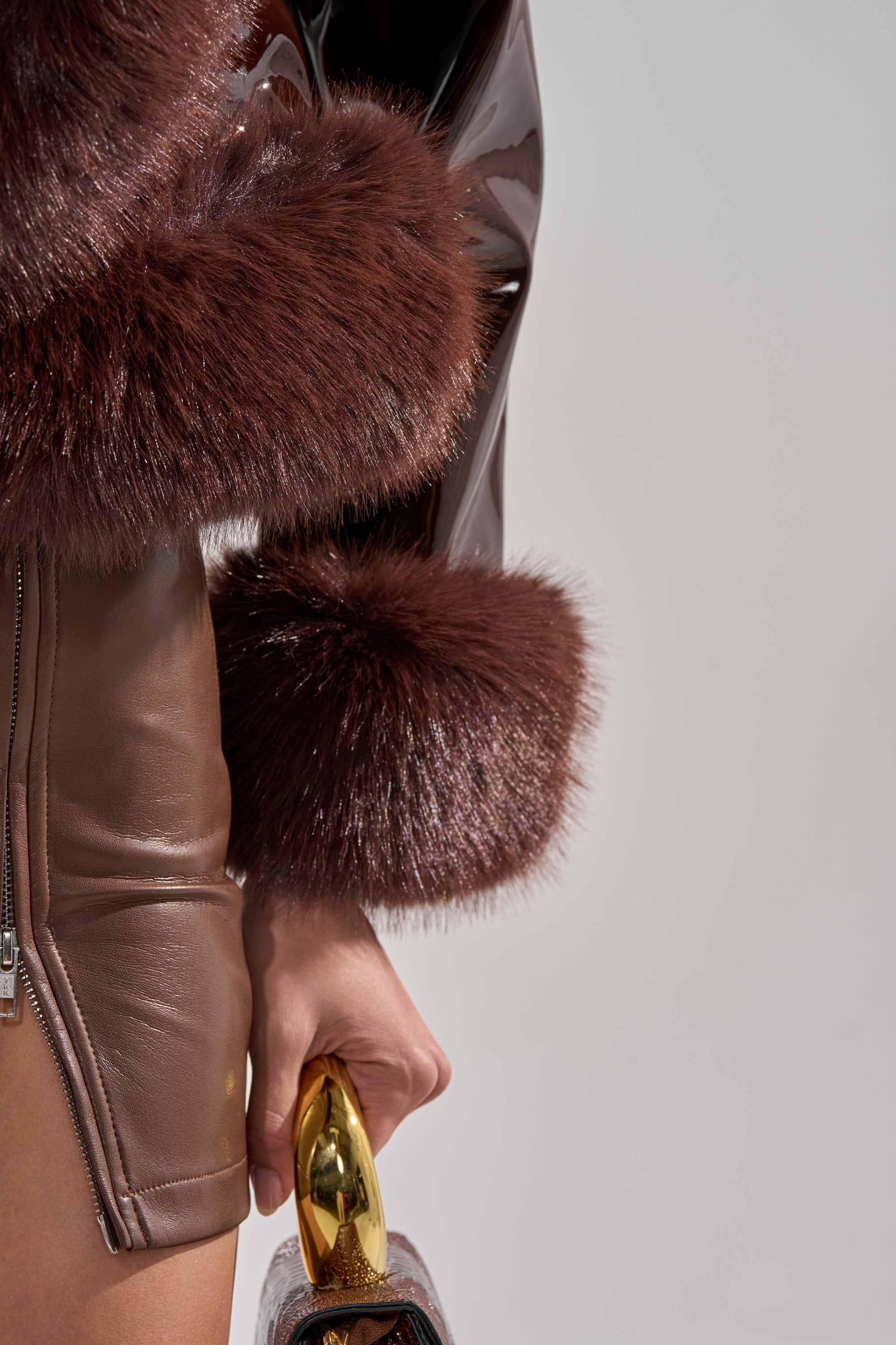 A person wears the LUNNA FUR TRIM MOTO IN PATENT—a shiny brown faux leather skirt and matching cropped jacket with brown faux fur cuffs—holding a gold-handled handbag against a plain, neutral background.