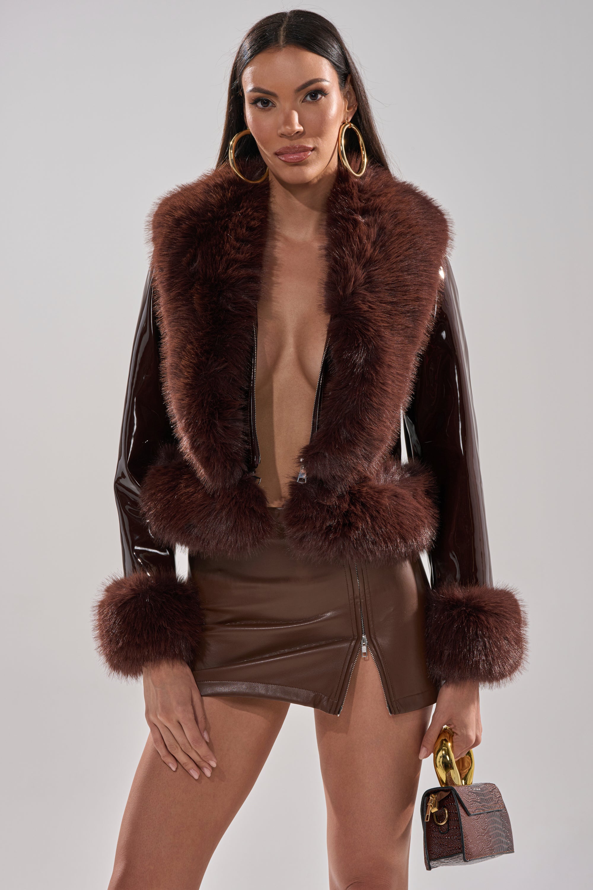 A woman wears the LUNNA FUR TRIM MOTO IN PATENT jacket with a matching mini skirt, carries a textured handbag, and accessorizes with large hoop earrings. Her long straight hair and confident look stand out against a plain light background.