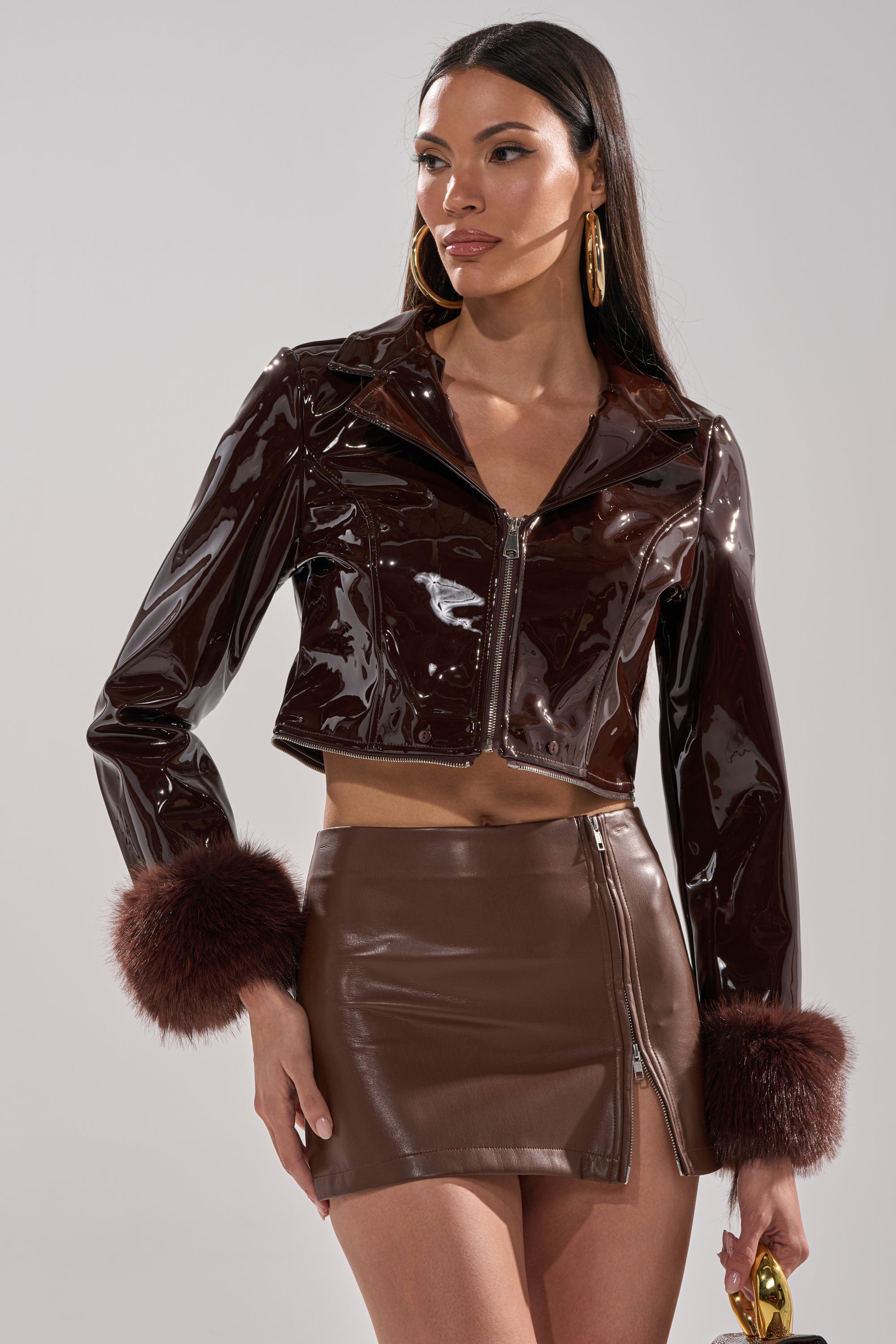 A woman wears the LUNNA FUR TRIM MOTO IN PATENT, a shiny brown faux leather jacket with fur cuffs, paired with a matching brown mini skirt with an angled zipper. She accessorizes with large hoop earrings and holds a small gold item.