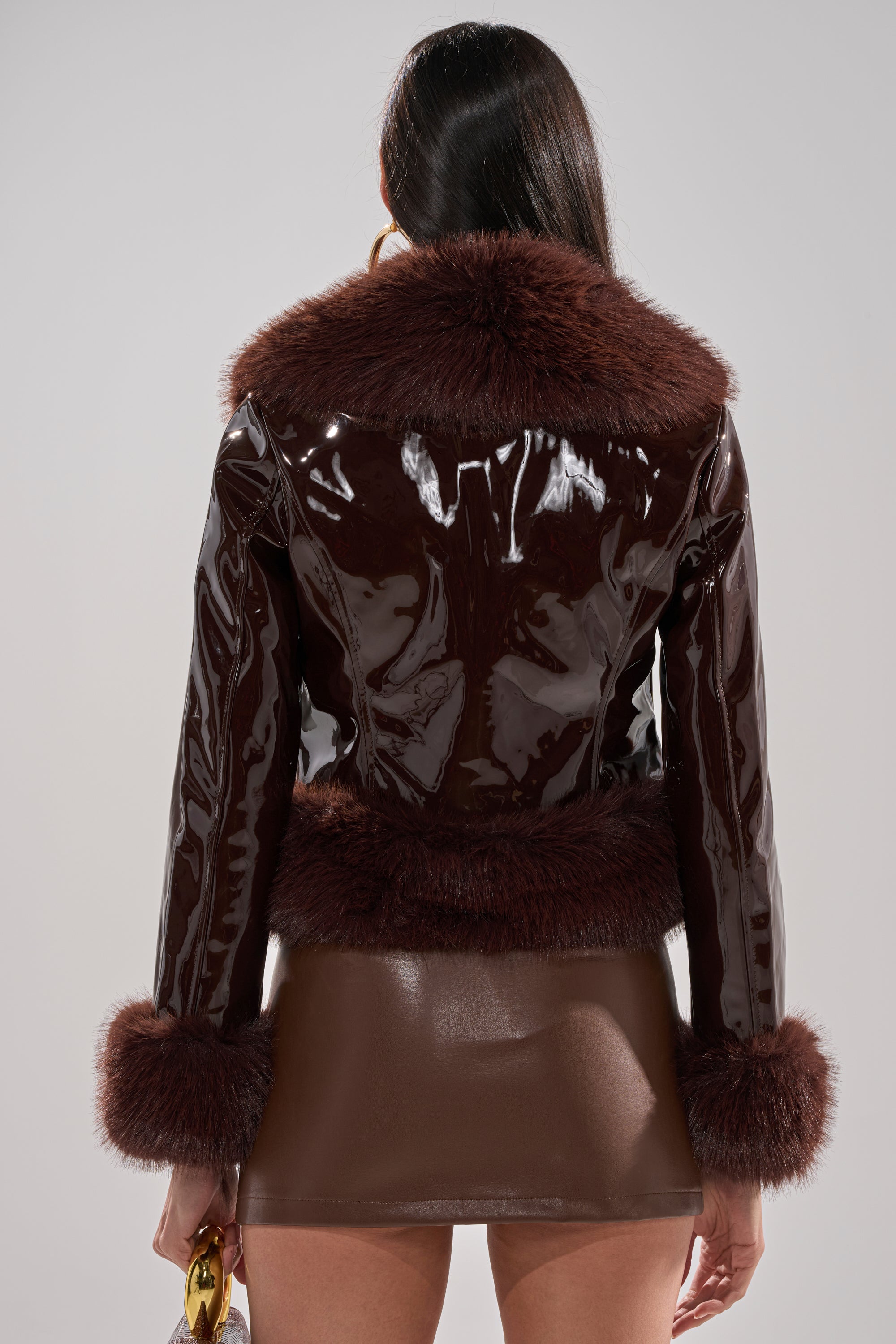 A person with long dark hair is seen from the back wearing the LUNNA FUR TRIM MOTO IN PATENT—a shiny dark brown jacket with faux fur collar, cuffs, and hem—paired with a brown mini skirt, holding a gold accessory.