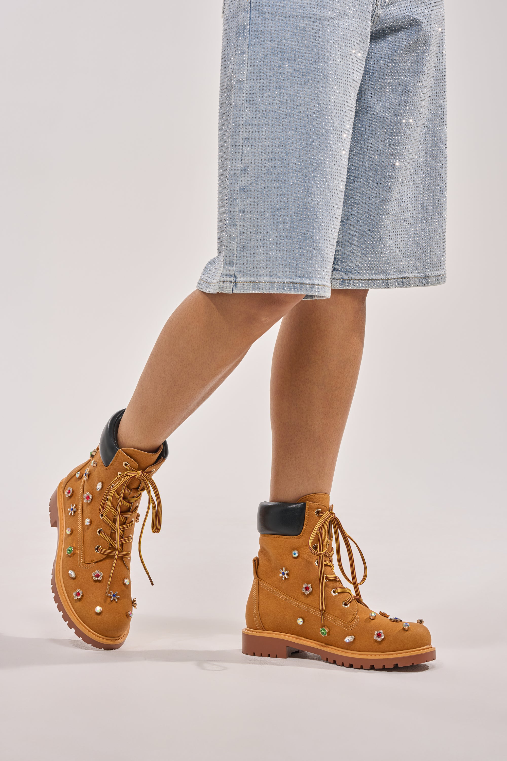 Wearing the AKIRA GOLD LUSTER TAN EMBELLISHED FLAT COMBAT FESTIVAL BOOTIE, a person pairs these flower-adorned boots with light blue, knee-length denim shorts for an effortless festival streetwear look against a light background. 0