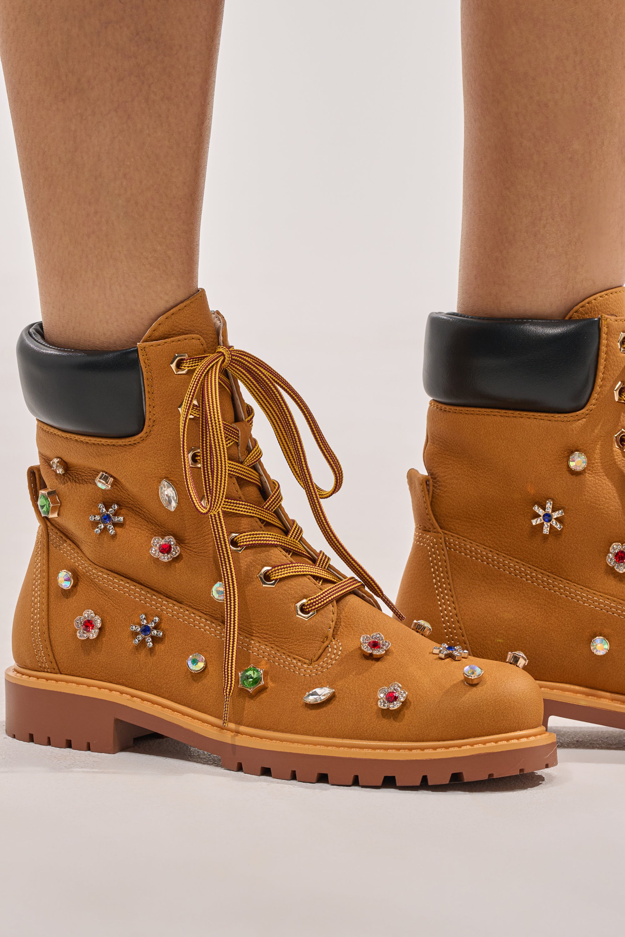 Close-up of the AKIRA GOLD LUSTER TAN EMBELLISHED FLAT COMBAT FESTIVAL BOOTIE featuring tan faux leather, colorful rhinestone and flower embellishments, black padded collars, thick treaded soles, and a flattering peek of lower legs. 3