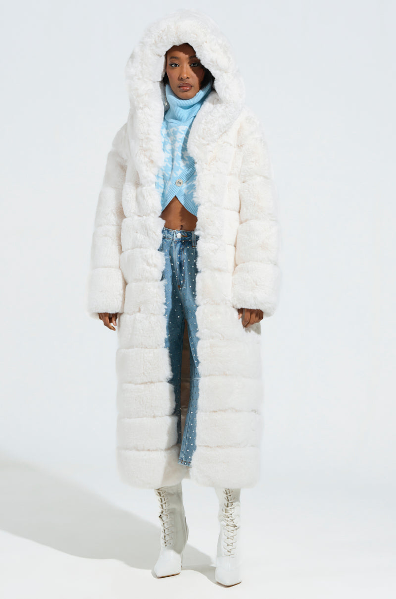 A person wearing the LUXE JENNY HOODED FAUX FUR TRENCH IN WHITE stands on a white background. Underneath, they sport a light blue ribbed turtleneck and matching wide-leg pants with a cutout design. White lace-up boots complete the look, epitomizing AKIRA Label's chic aesthetic.