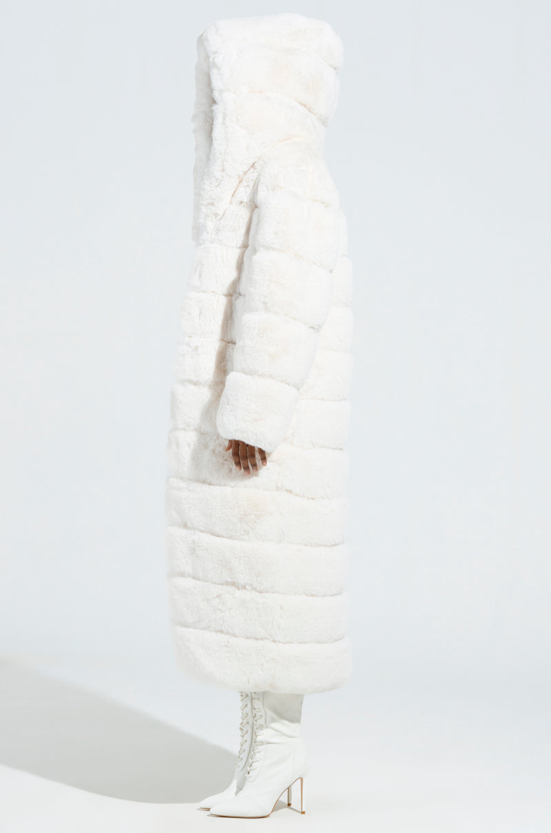 Against a plain white backdrop, the model is luxuriously draped in the LUXE JENNY HOODED FAUX FUR TRENCH IN WHITE, complemented by elegant white heeled boots. This AKIRA Label creation showcases a quilted pattern and elegantly reaches mid-calf, exuding sophistication even as the model's face remains hidden.