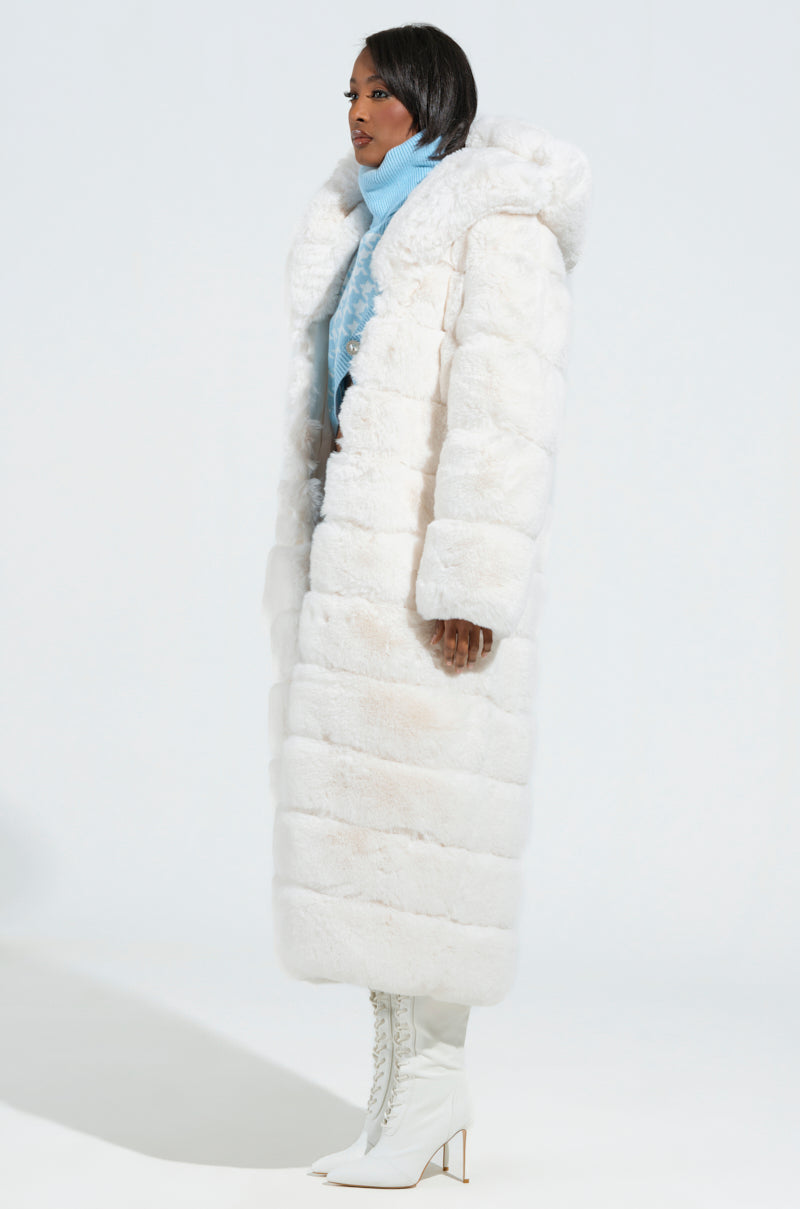 Wearing the Luxe Jenny Hooded Faux Fur Trench in white and white high-heeled boots, a person stands against a light background. The light blue turtleneck sweater underneath adds to their elegance as they gaze to the side.