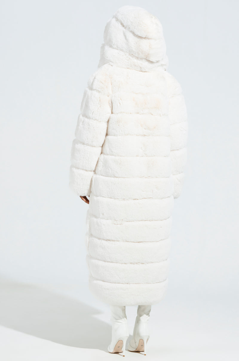Dressed in the AKIRA Label's Luxe Jenny Hooded Faux Fur Trench in White, an individual stands with their back facing the viewer. The coat's length extends to the ankles, complemented by coordinating white shoes. The backdrop is a simple, light hue.