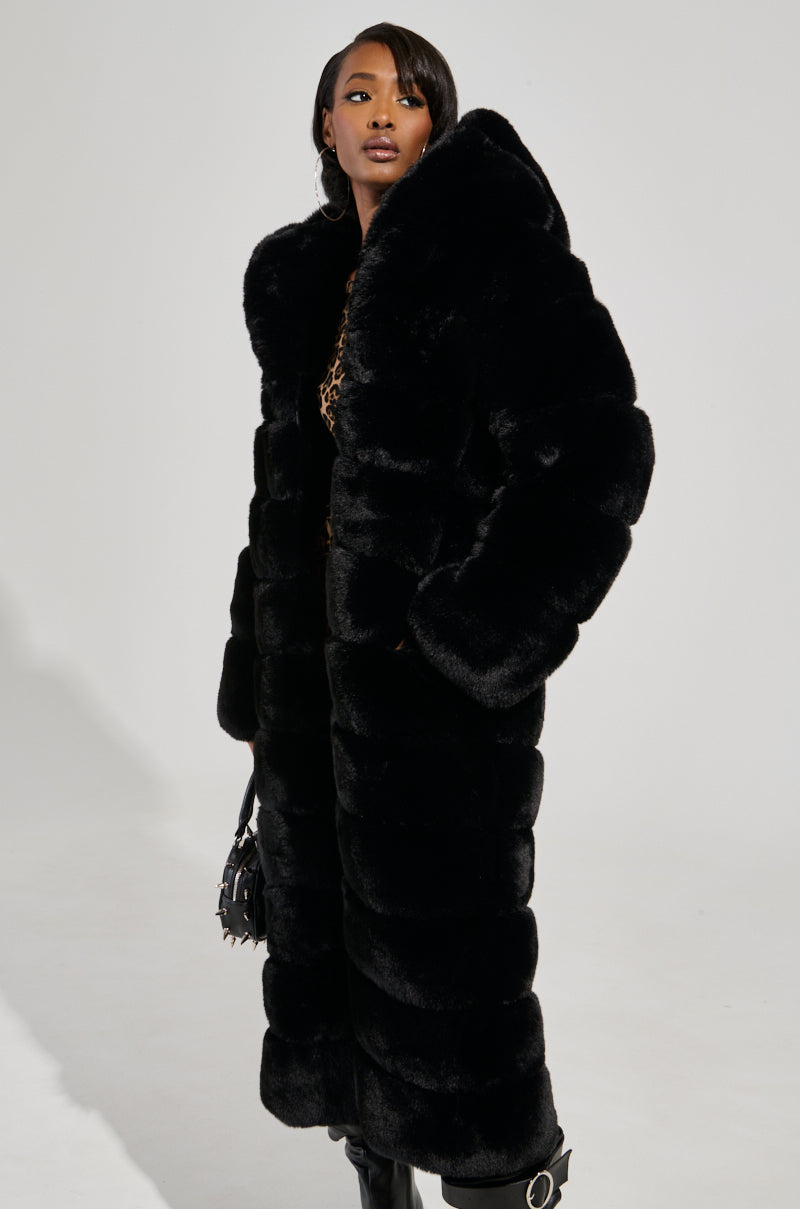 Wearing the FAUX FUR PANEL HOODED TRENCH IN BLACK, a person stands confidently, clutching a small black handbag. Their long hair is elegantly adorned with a luxe headband, as they gaze to the side against a neutral background.