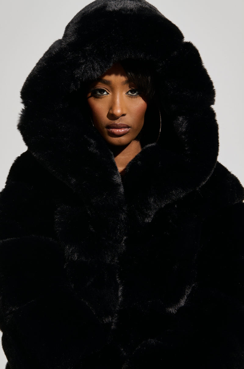 Wearing the FAUX FUR PANEL HOODED TRENCH IN BLACK with the hood up, a person looks directly at the camera with a serious expression, highlighting the coat’s plush elegance against a plain background.