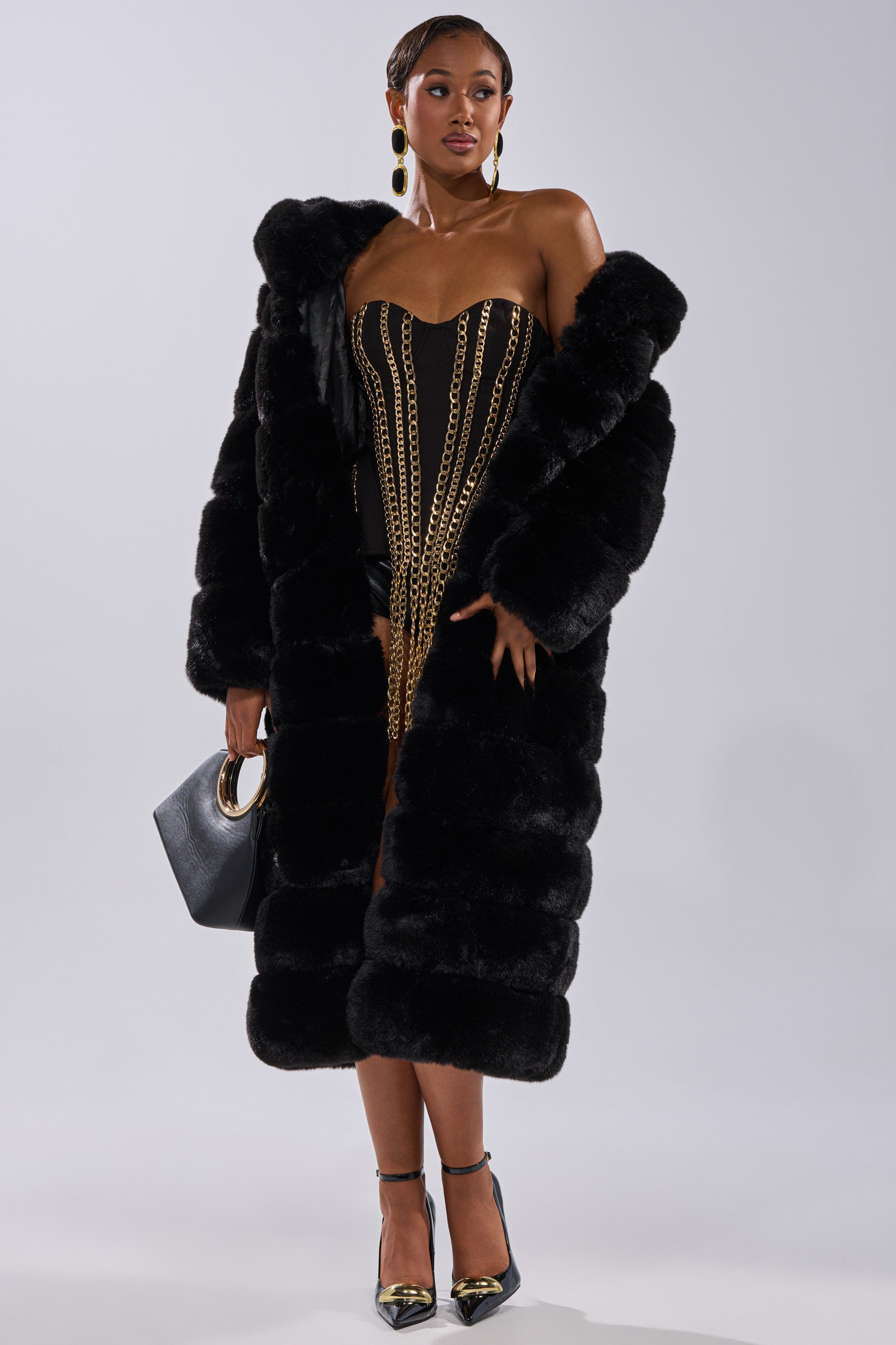 A woman stands confidently in the FAUX FUR PANEL HOODED TRENCH IN BLACK over a strapless black dress with gold chain details, black heels with gold tips, statement earrings, and a black handbag with a gold handle.