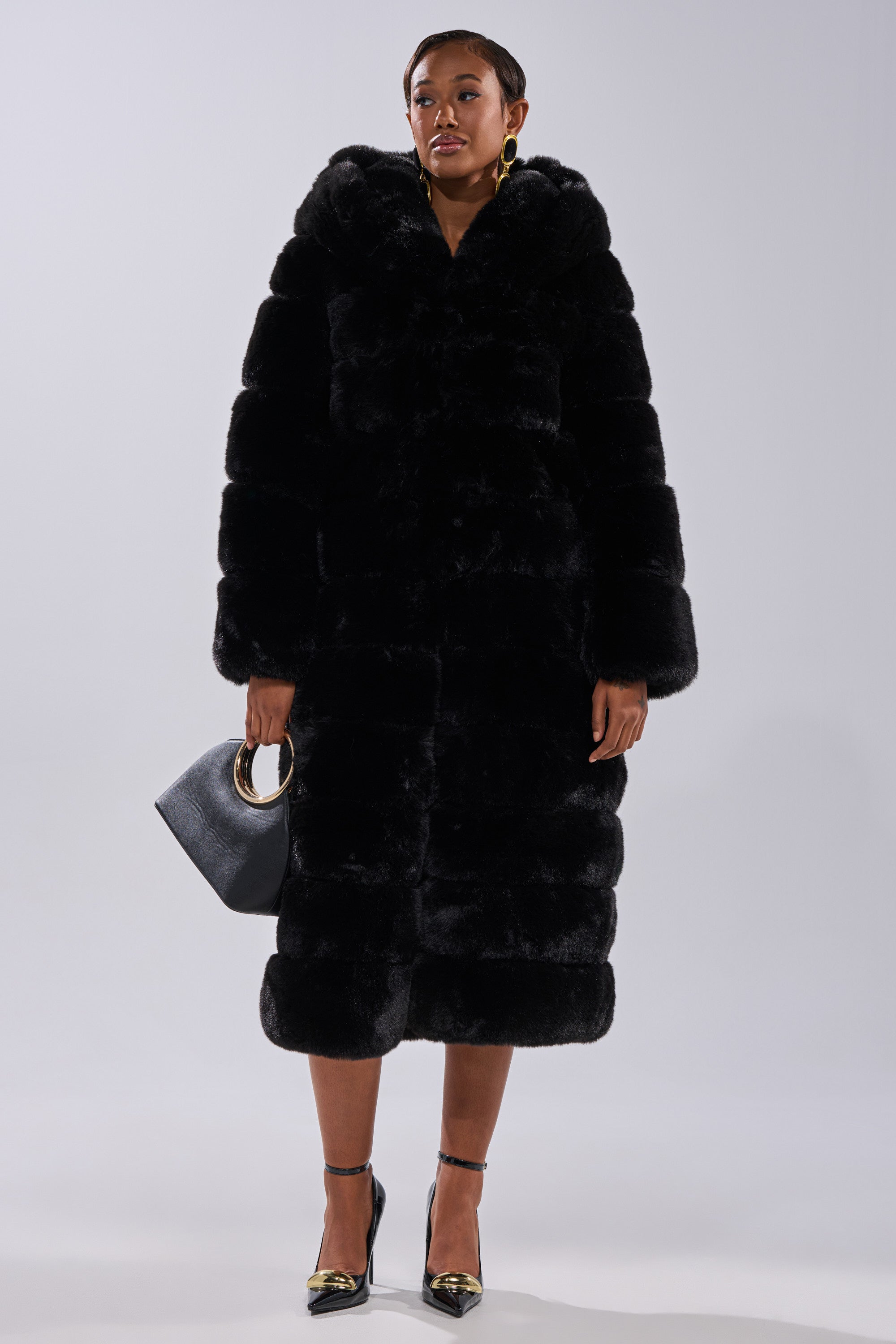 A person faces forward, wearing the FAUX FUR PANEL HOODED TRENCH IN BLACK, paired with a black handbag with gold handles, gold earrings, and black high heels. Their expression is neutral.
