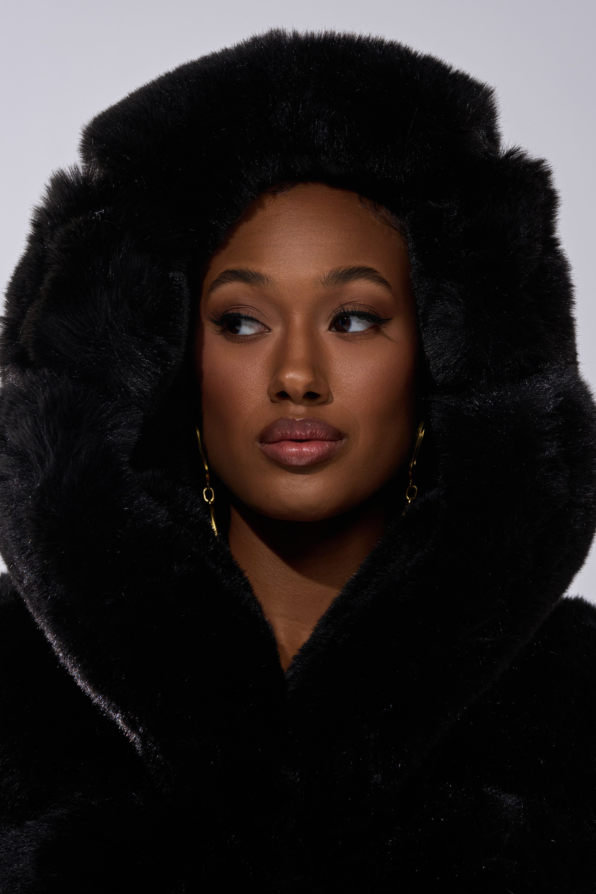A woman wearing the FAUX FUR PANEL HOODED TRENCH IN BLACK looks to the side, her face framed by its large, fluffy hood and gold dangling earrings. The background is plain and neutral.