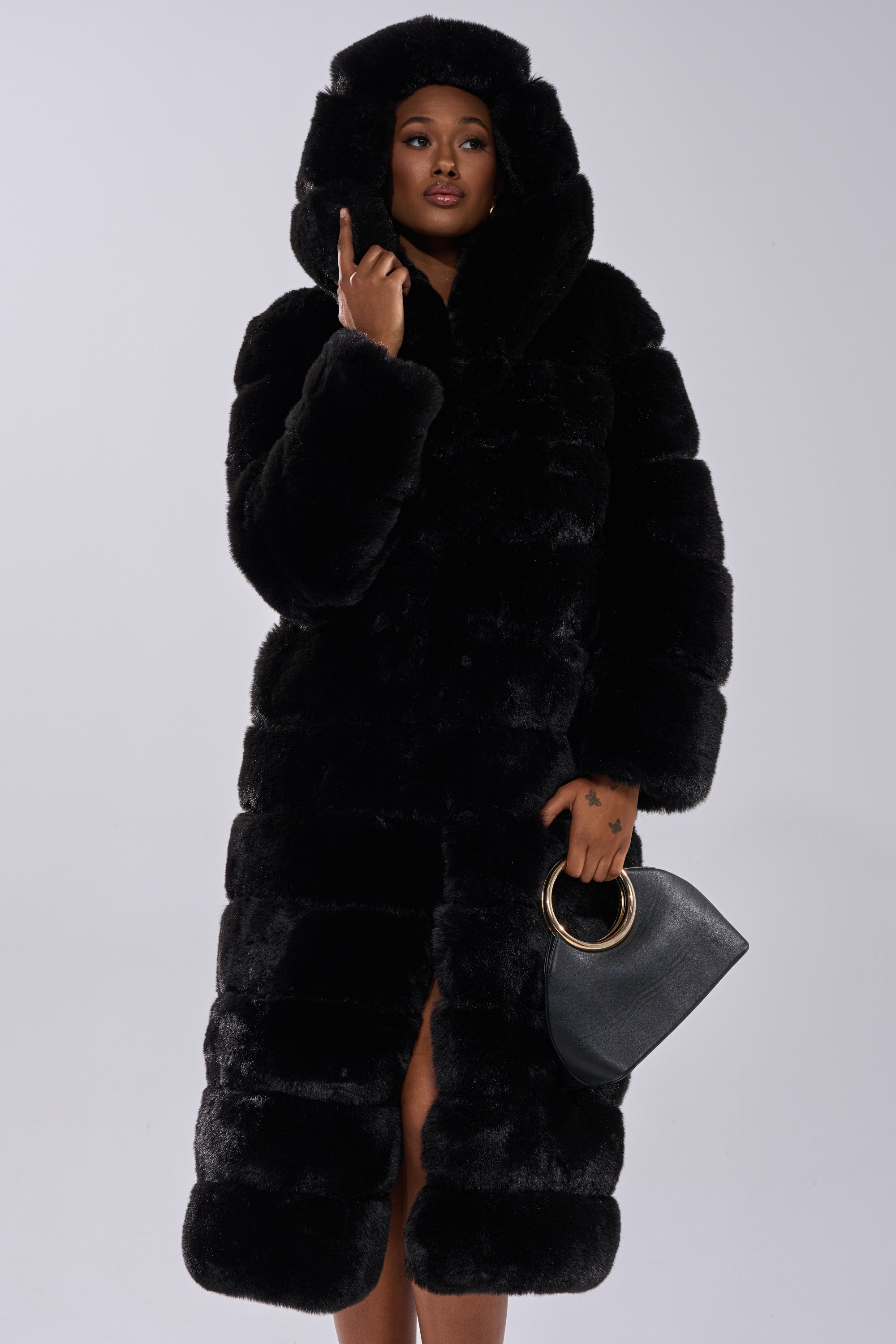 A person models the FAUX FUR PANEL HOODED TRENCH IN BLACK, standing confidently with a black handbag against a plain light background.