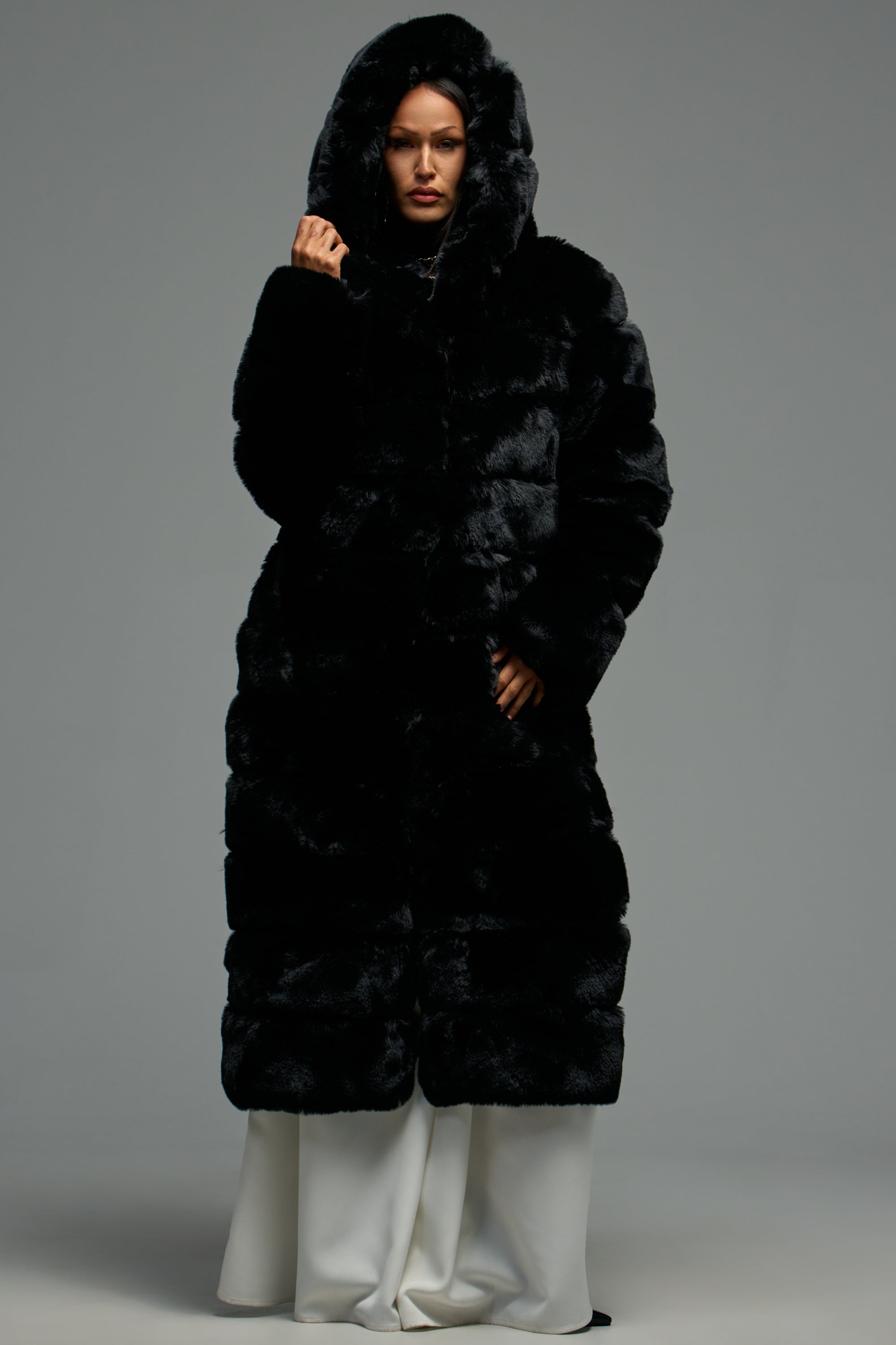 A person stands facing forward in the FAUX FUR PANEL HOODED TRENCH IN BLACK, styled with white pants against a plain gray background.