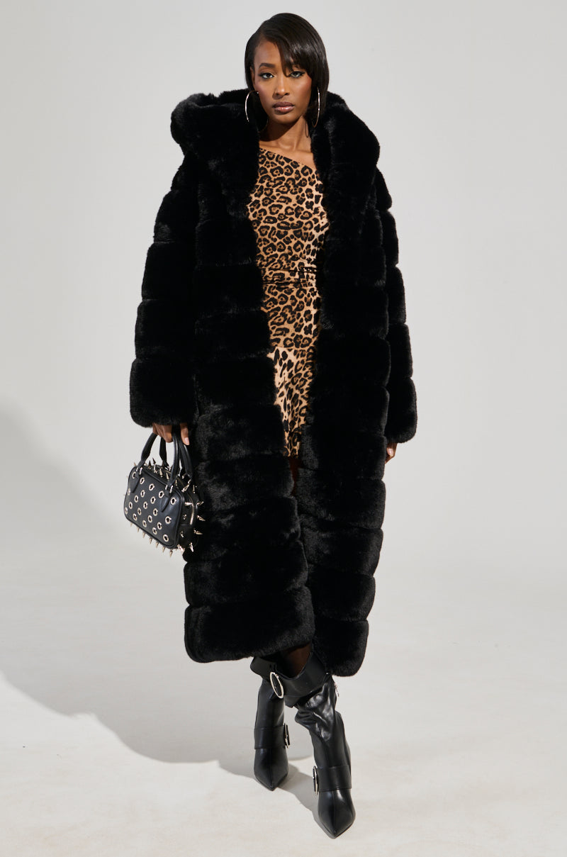 A woman stands against a plain light background wearing the FAUX FUR PANEL HOODED TRENCH IN BLACK over a leopard print dress, black knee-high boots with buckles, and holding a small black handbag with silver eyelet details.