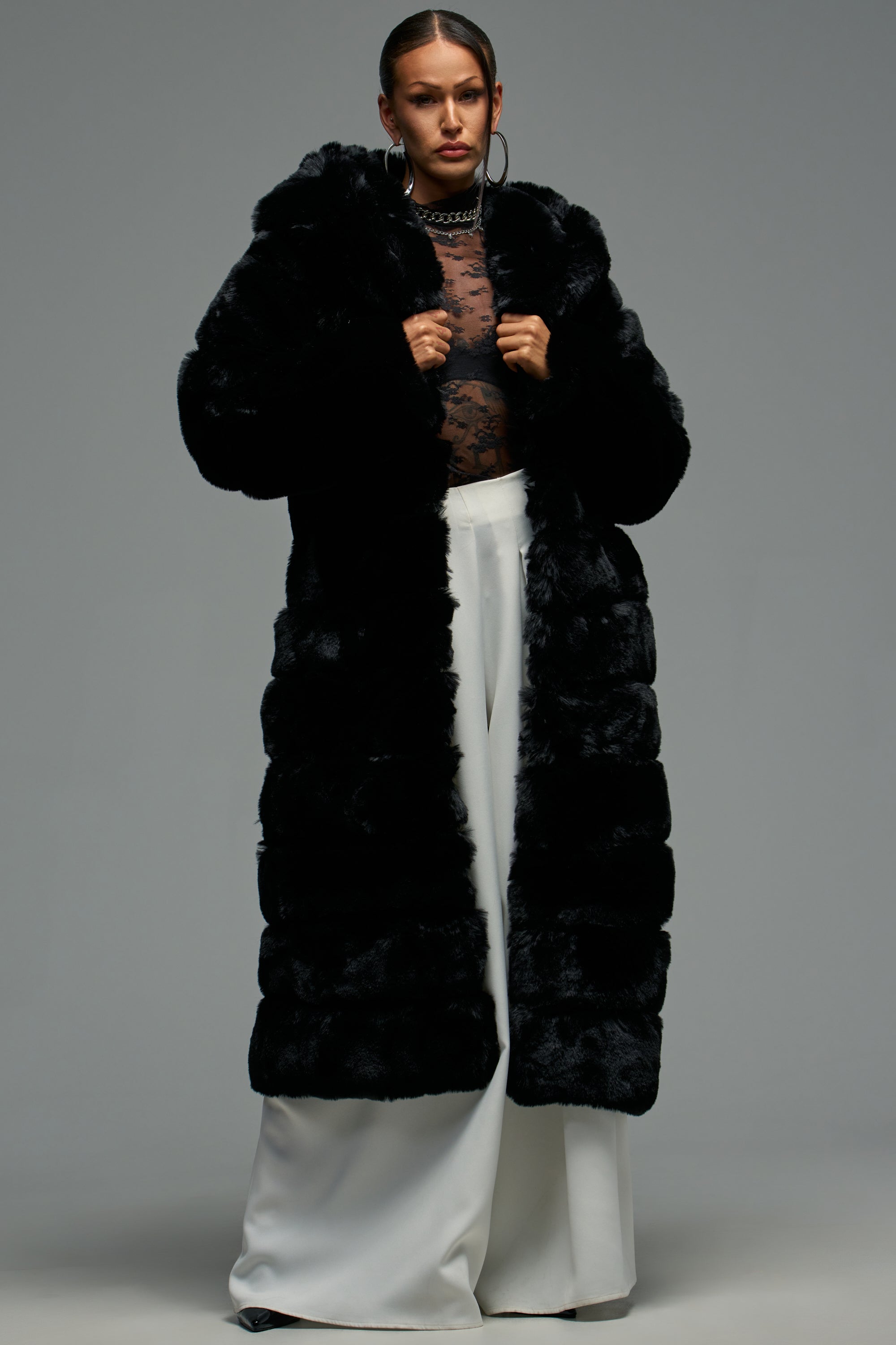 A woman stands confidently in the FAUX FUR PANEL HOODED TRENCH IN BLACK, styled with a sheer black lace top, white wide-leg pants, hoop earrings, and a chunky chain necklace against a plain gray background.