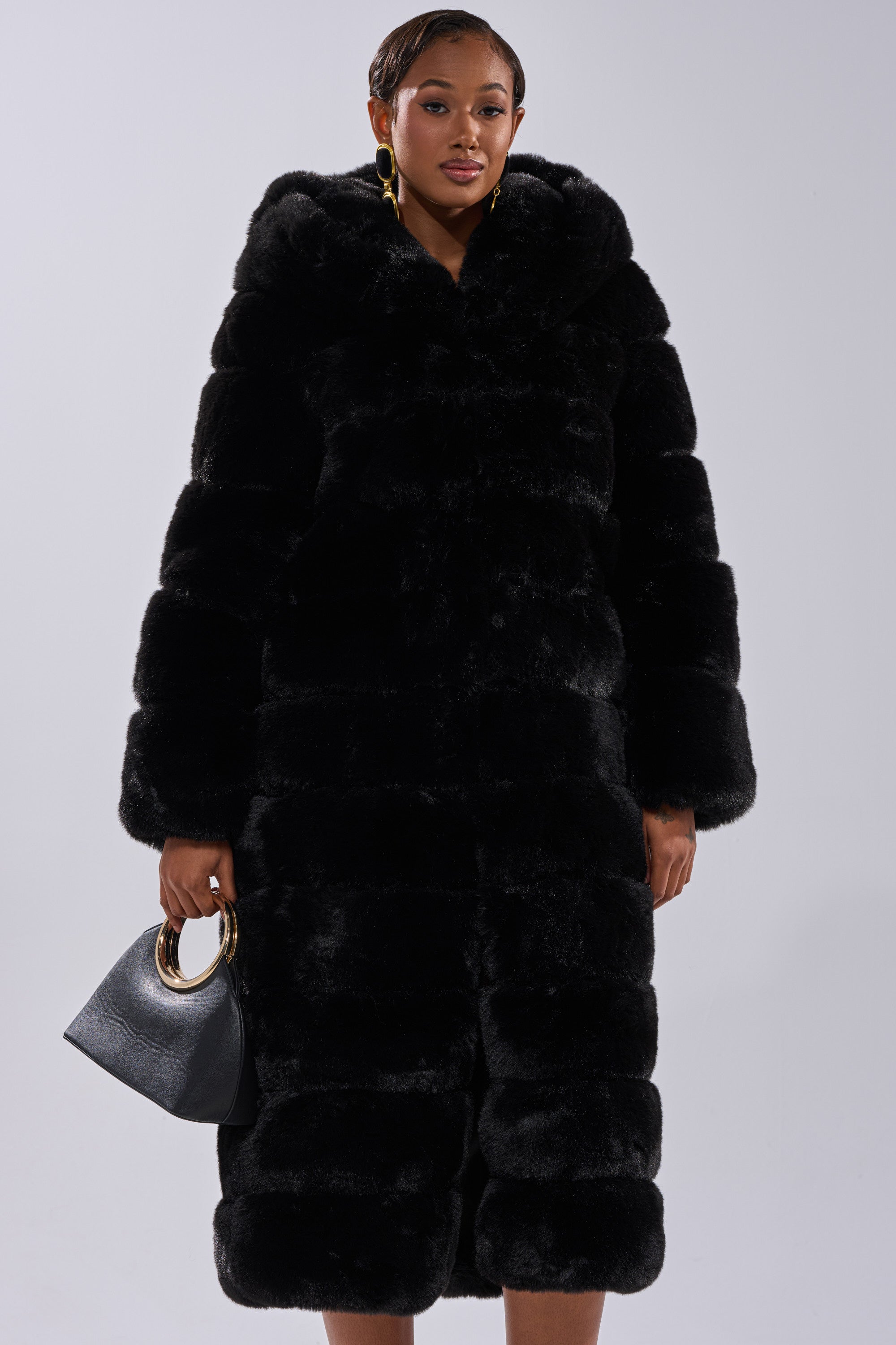 A woman stands against a white background wearing the FAUX FUR PANEL HOODED TRENCH IN BLACK, a long, plush black faux fur coat with a hood, and holds a black handbag with gold handles.