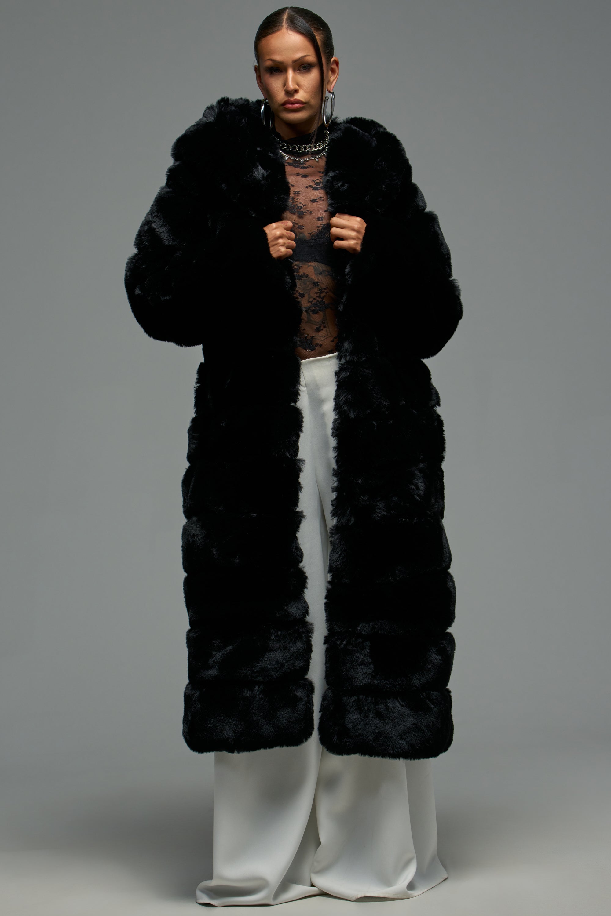 A person wears the FAUX FUR PANEL HOODED TRENCH IN BLACK over a sheer black lace top and wide-legged white pants, standing confidently against a plain grey background.