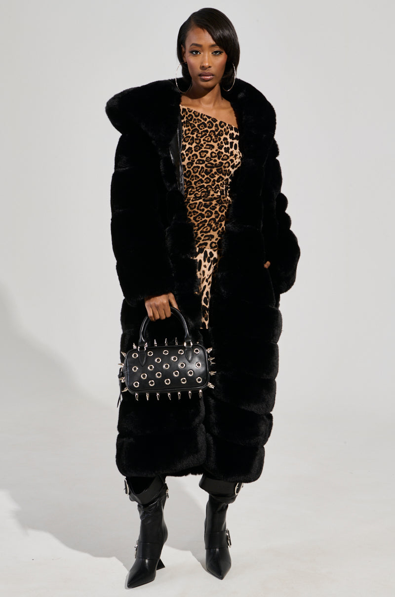 A woman stands confidently in the FAUX FUR PANEL HOODED TRENCH IN BLACK over a one-shoulder leopard print dress, accessorizing with a studded black handbag and black pointed boots with buckles.