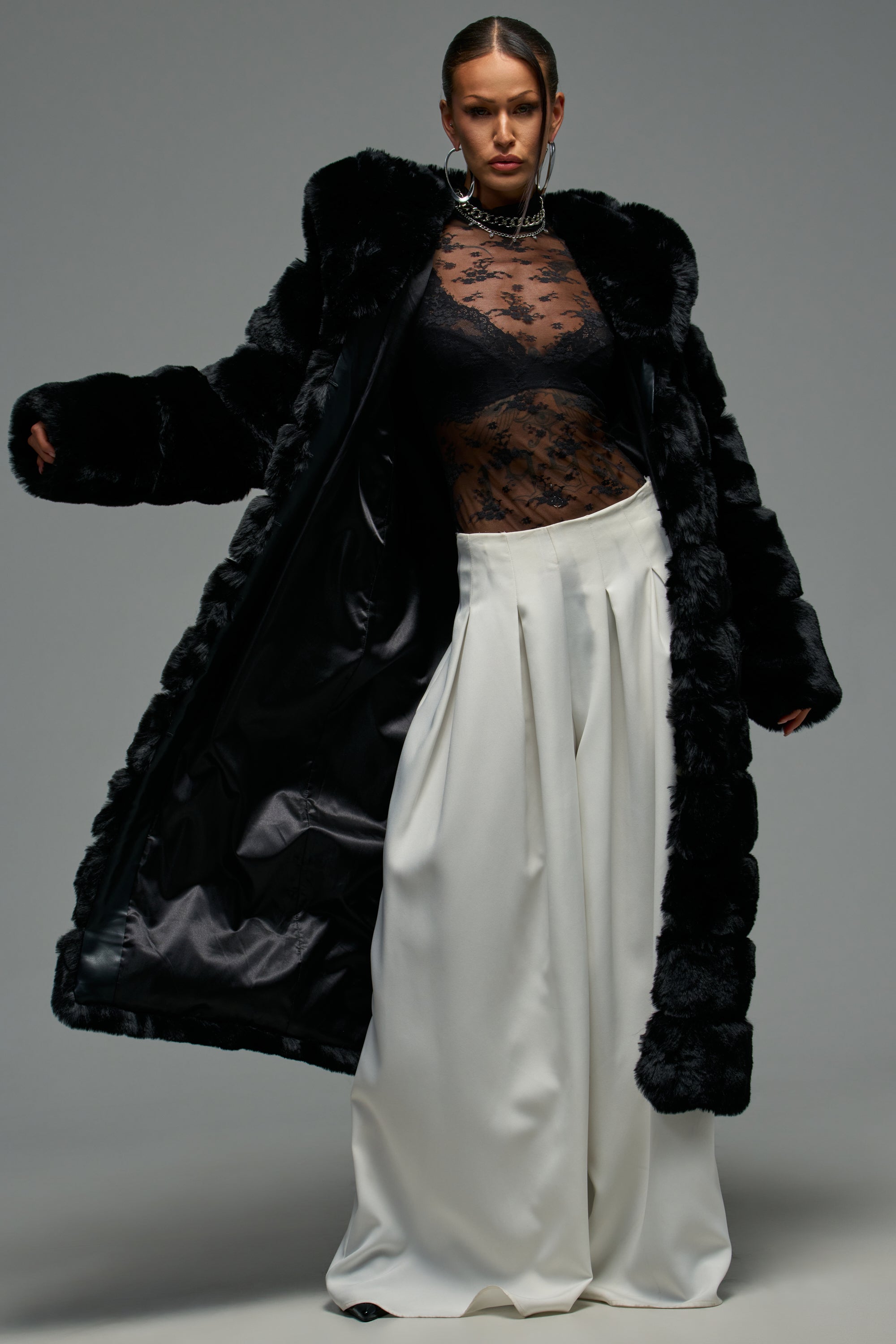A woman models the FAUX FUR PANEL HOODED TRENCH IN BLACK over a sheer black lace top and wide-leg white pants, standing against a plain gray background.