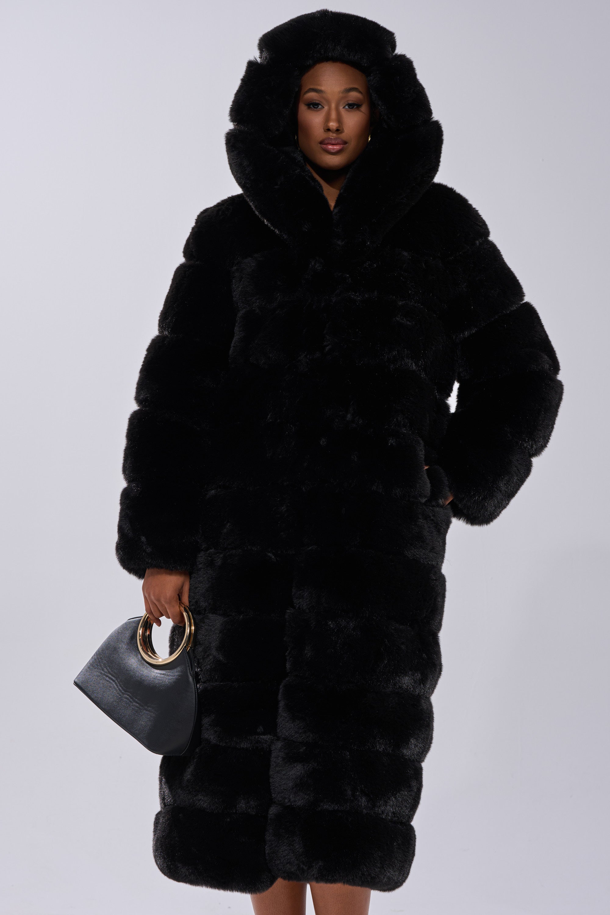A woman wearing the FAUX FUR PANEL HOODED TRENCH IN BLACK stands against a plain background, holding a black bag with gold handles in her left hand.