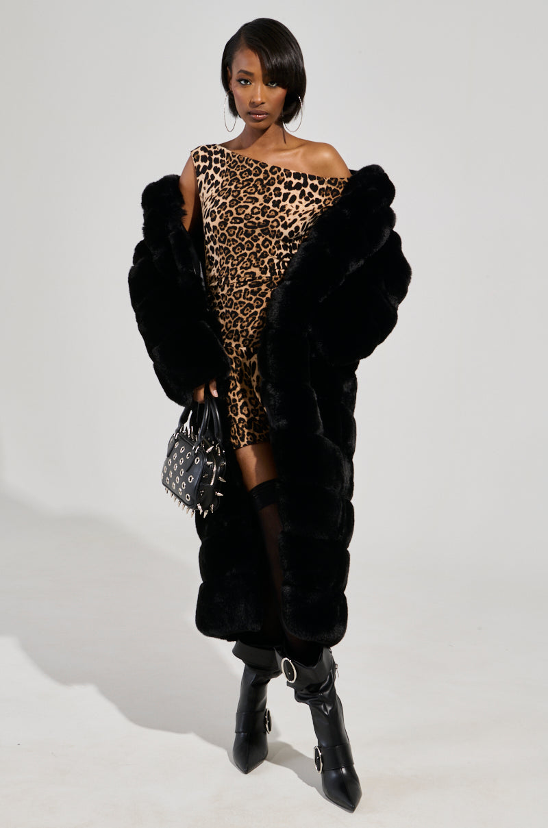 A woman stands confidently in a one-shoulder leopard print dress, black heeled boots with buckles, a black studded handbag, and the FAUX FUR PANEL HOODED TRENCH IN BLACK against a plain light background.