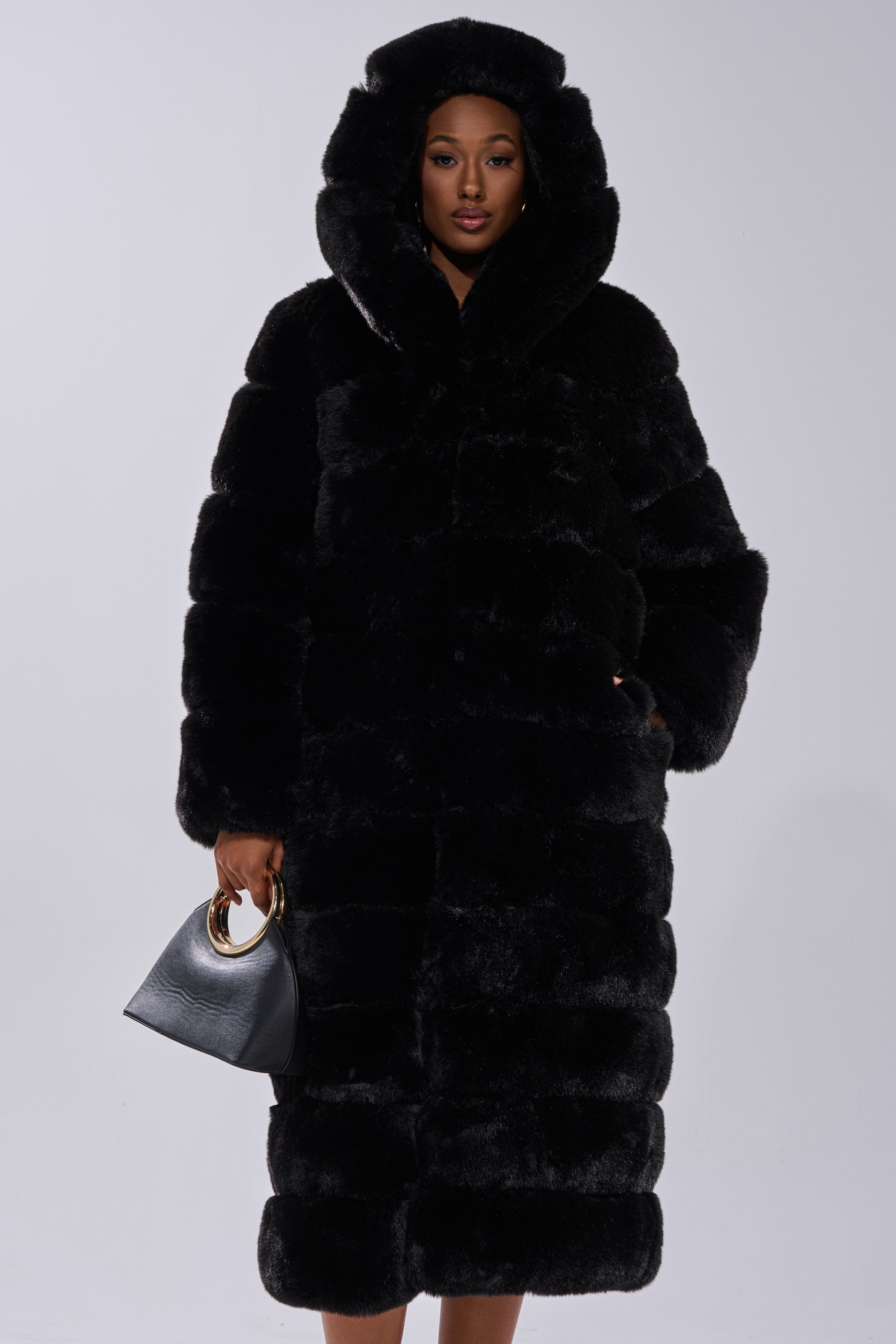 A person stands against a plain background wearing the FAUX FUR PANEL HOODED TRENCH IN BLACK, paired with a black handbag with gold handles in their left hand.