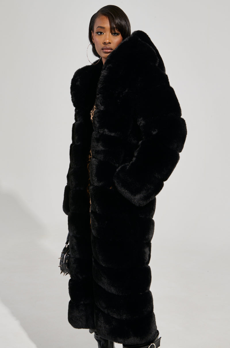 A woman stands confidently in the FAUX FUR PANEL HOODED TRENCH IN BLACK, styled with black boots and hoop earrings, against a plain light gray background.