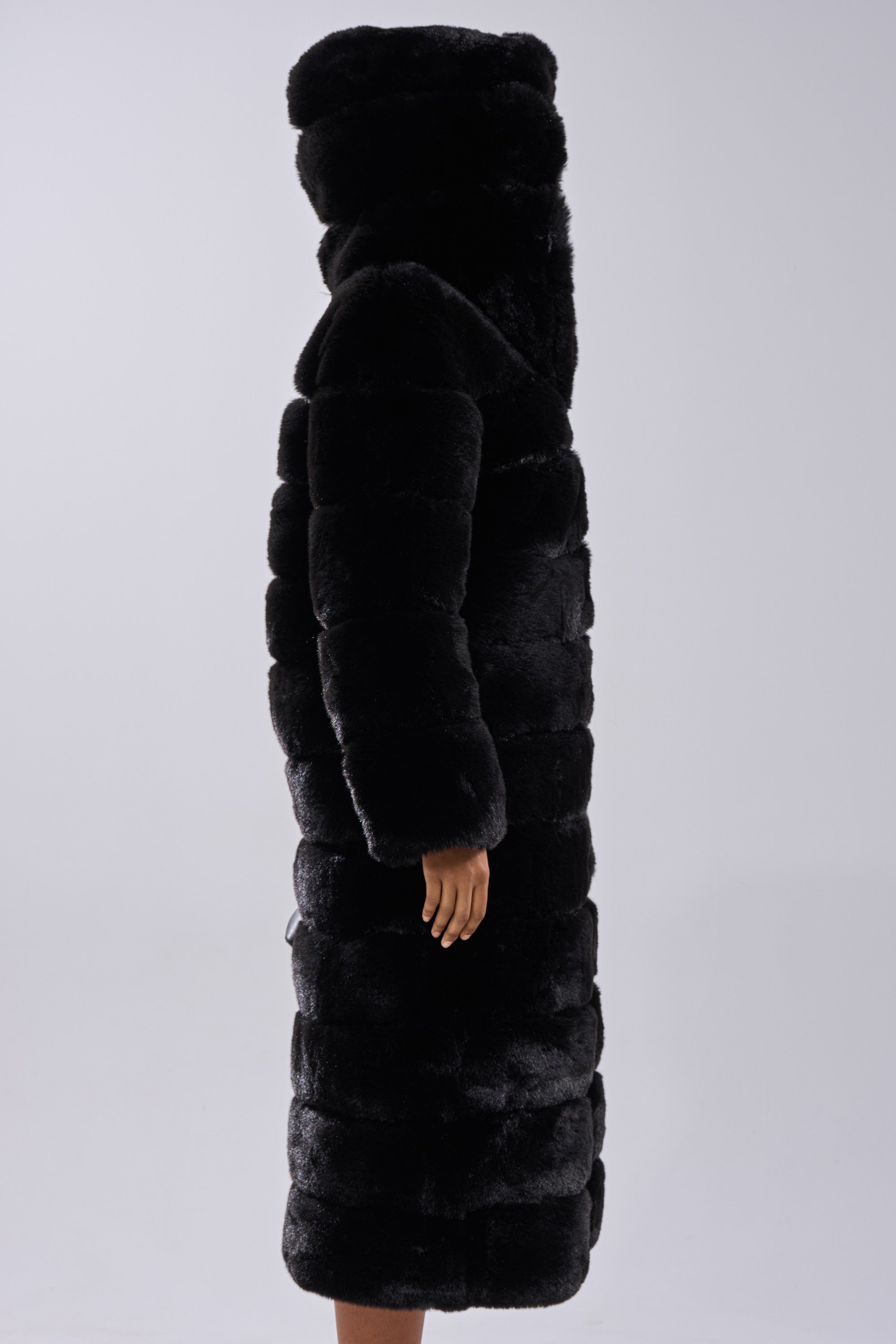 A person stands sideways against a light gray background, wearing the FAUX FUR PANEL HOODED TRENCH IN BLACK; only their hand and part of their face are visible.