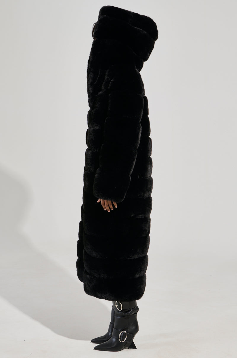 A person stands in profile wearing the FAUX FUR PANEL HOODED TRENCH IN BLACK, featuring a long length and large hood, paired with black pointed boots with silver buckles against a plain light background.