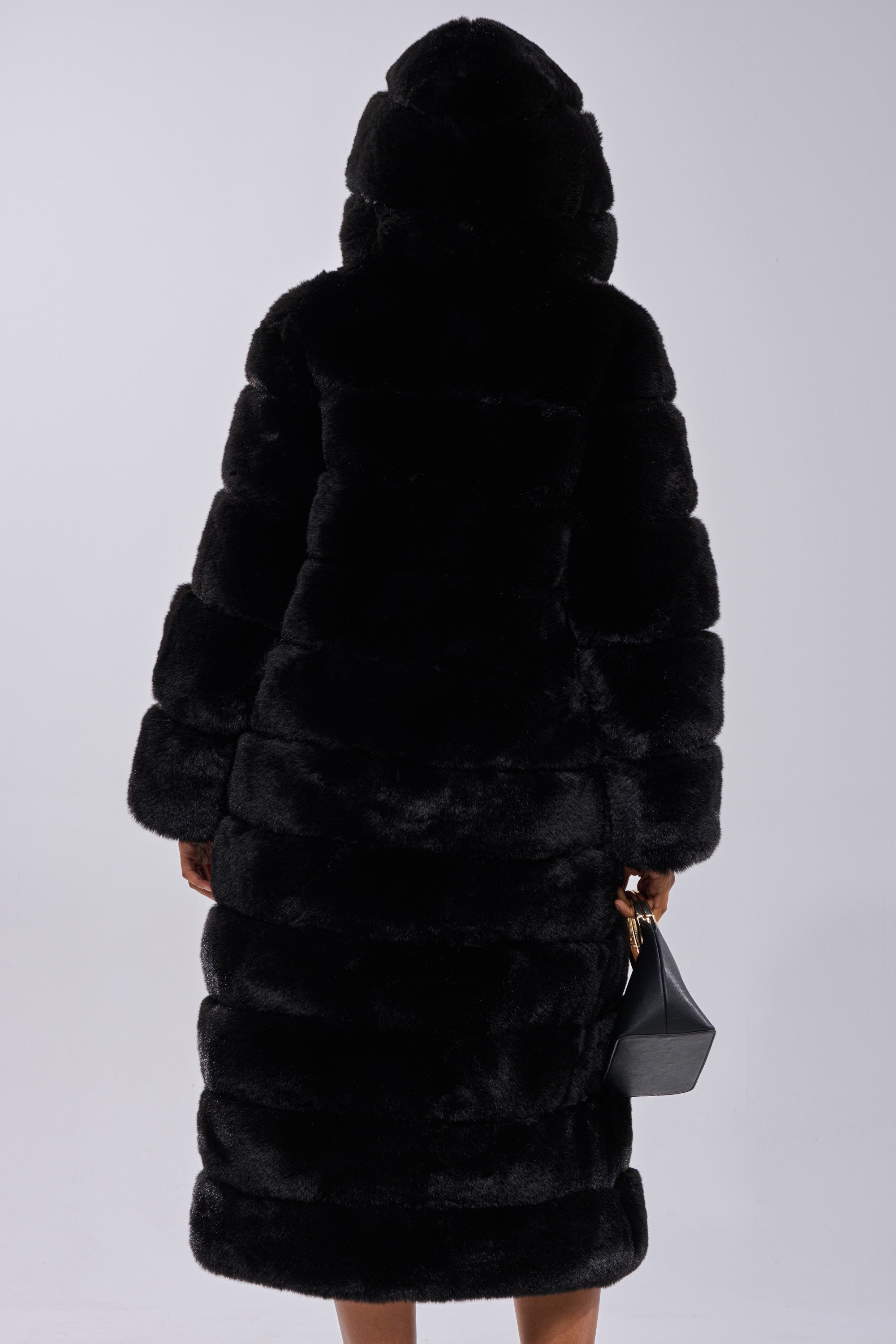 A person faces away, wearing the FAUX FUR PANEL HOODED TRENCH IN BLACK and holding a small black handbag in their left hand, set against a plain light background.