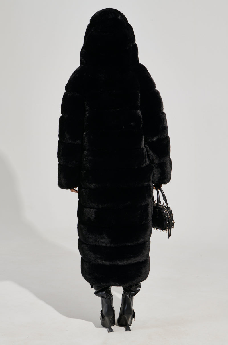 Wearing the FAUX FUR PANEL HOODED TRENCH IN BLACK, a person stands facing away, holding a small handbag and wearing tall black boots against a plain, light-colored background.