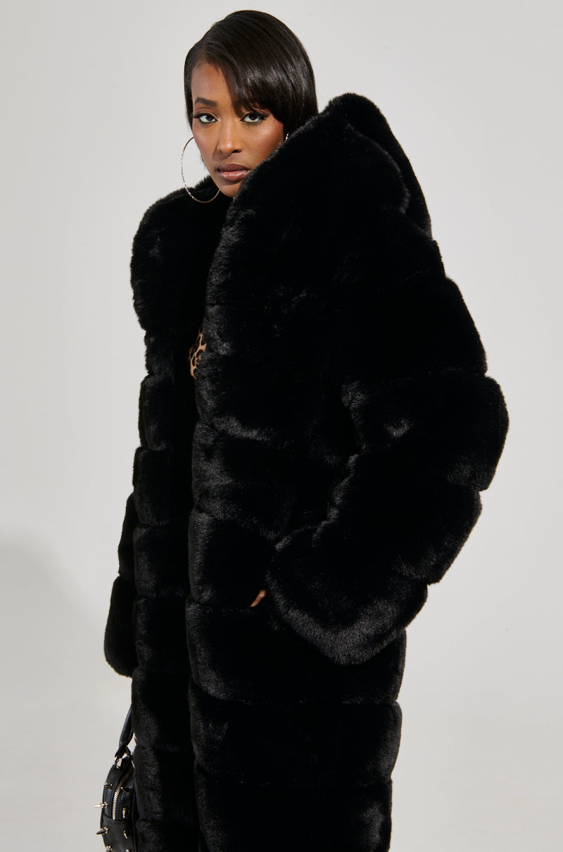 A woman models the FAUX FUR PANEL HOODED TRENCH IN BLACK, an oversized faux fur coat with a hood, against a light background. She pairs it with hoop earrings and a sleek hairstyle, exuding confidence as she looks at the camera.