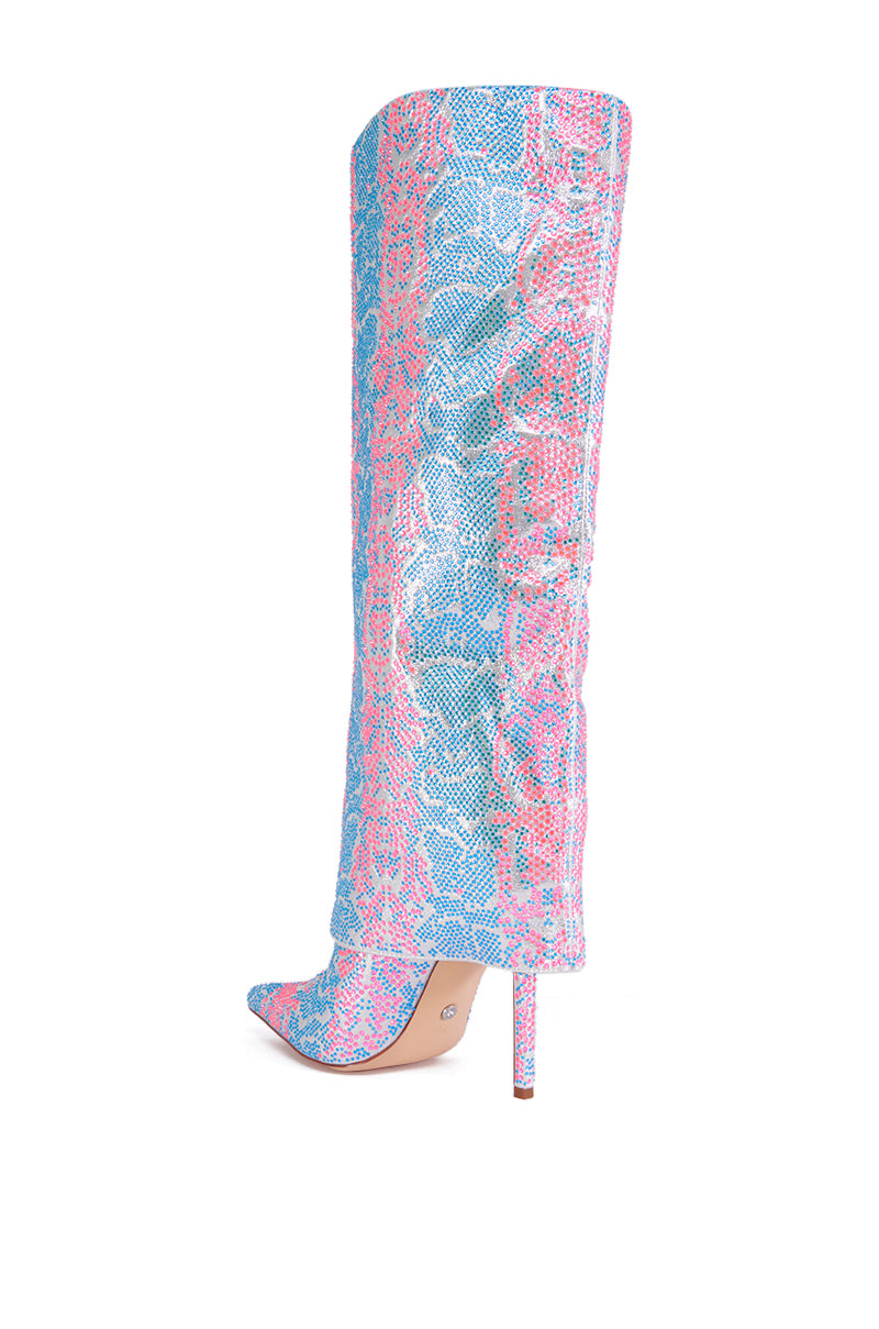A close-up view of the AZALEA WANG LUXER PINK AND BLUE MULTI RHINESTONE BOOT, showcasing its tall, high-heeled design with a pointed toe. The boot features a vibrant snake print pattern in blue, pink, and purple on a white background. It has a thin stiletto heel in light beige color. Bold and stylish, the design flaunts the flair of a glittery textile upper.