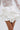 A person wearing a white lace long-sleeve top and the LUX WAVES MINI SKIRT, a high-waist sculptural ruffled mini skirt. The image highlights the detailed lace and the dramatic, wavy design of the skirt.