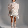 A woman stands in a studio wearing the LUX WAVES MINI SKIRT, a chic high-waist mini skirt, paired with a white lace top featuring an oversized bow, lace gloves, and tall white platform boots. Her hair is sleek and her expression neutral.