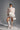 A woman poses in a high-fashion outfit featuring a sheer lace top with an oversized bow, the LUX WAVES MINI SKIRT with wavy ruffles and a high waist, tall white platform boots with cutouts, and statement earrings against a plain gray background.