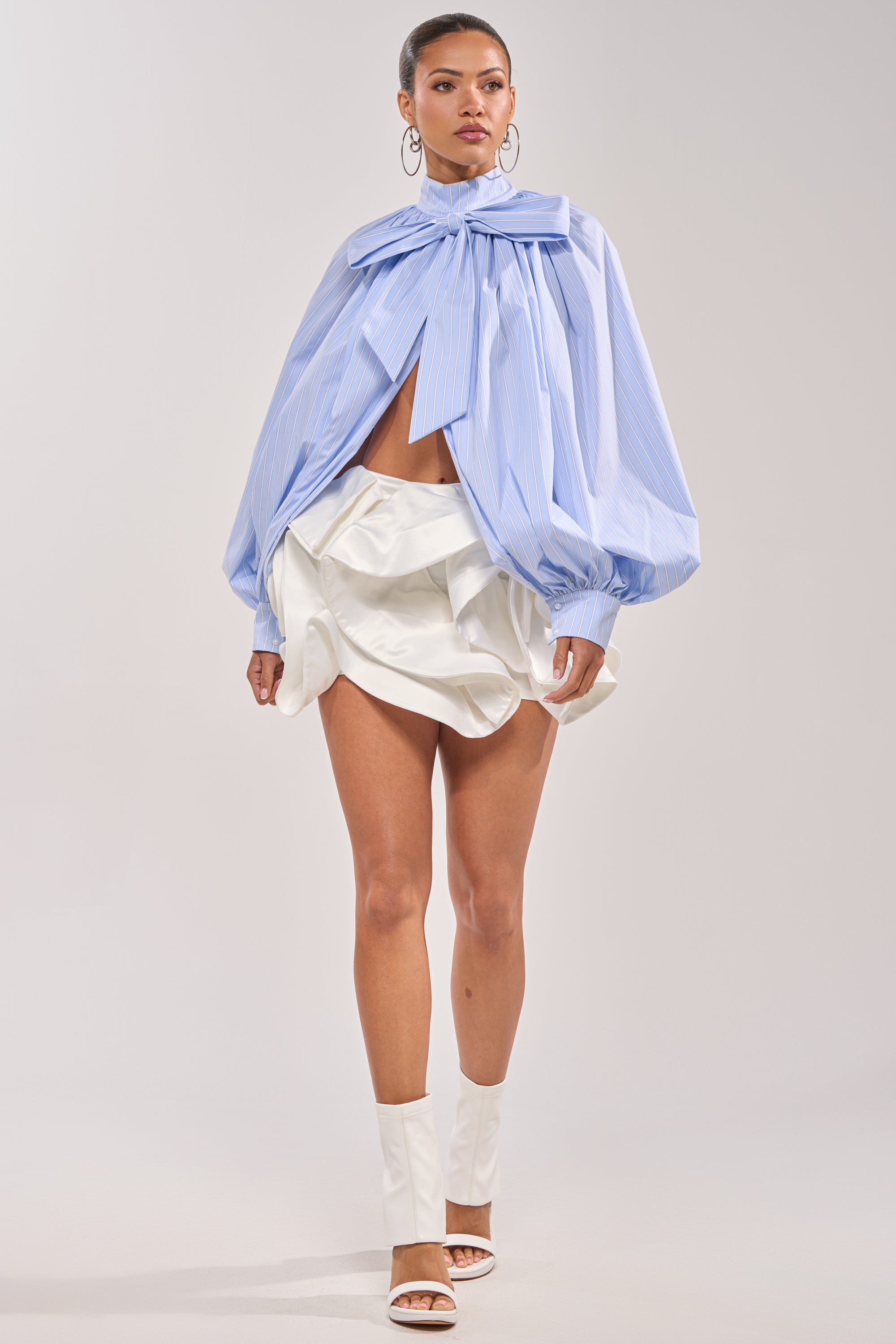 A woman models the LUX WAVES MINI SKIRT, styled with a light blue oversized bow-tie blouse, voluminous sleeves, and white open-toe high-heeled boots against a plain background. 5