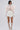 A woman stands against a white background in a sheer lace top with oversized pale peach bows, lace gloves, knee-high white gladiator boots, and the LUX WAVES MINI SKIRT—a white mini skirt featuring a wavy ruffle design. Her hair is sleek and wavy.